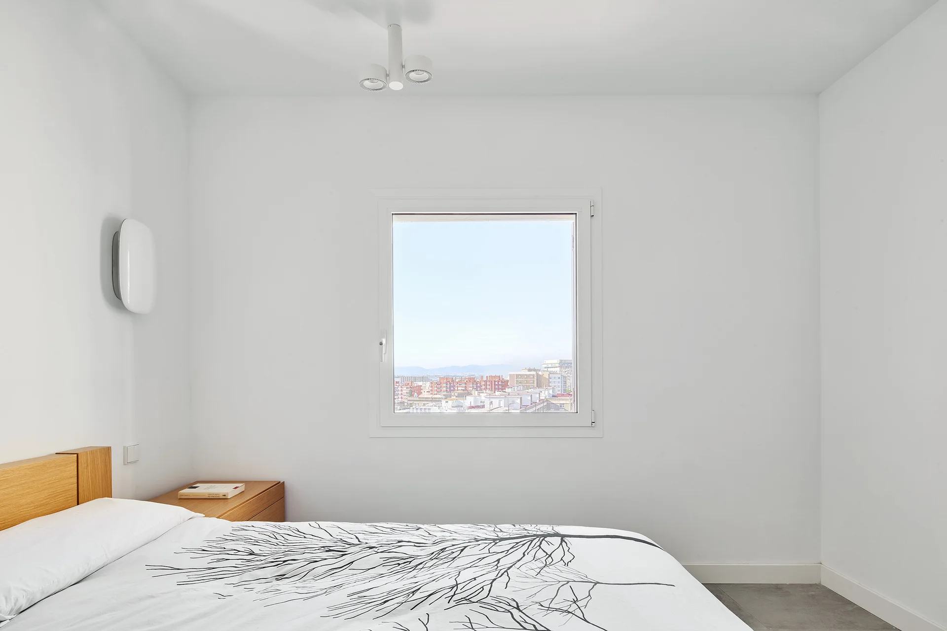 A minimalist bedroom features white walls, a wood headboard and nightstand, and a bed with a white-and-black tree branch patterned duvet.