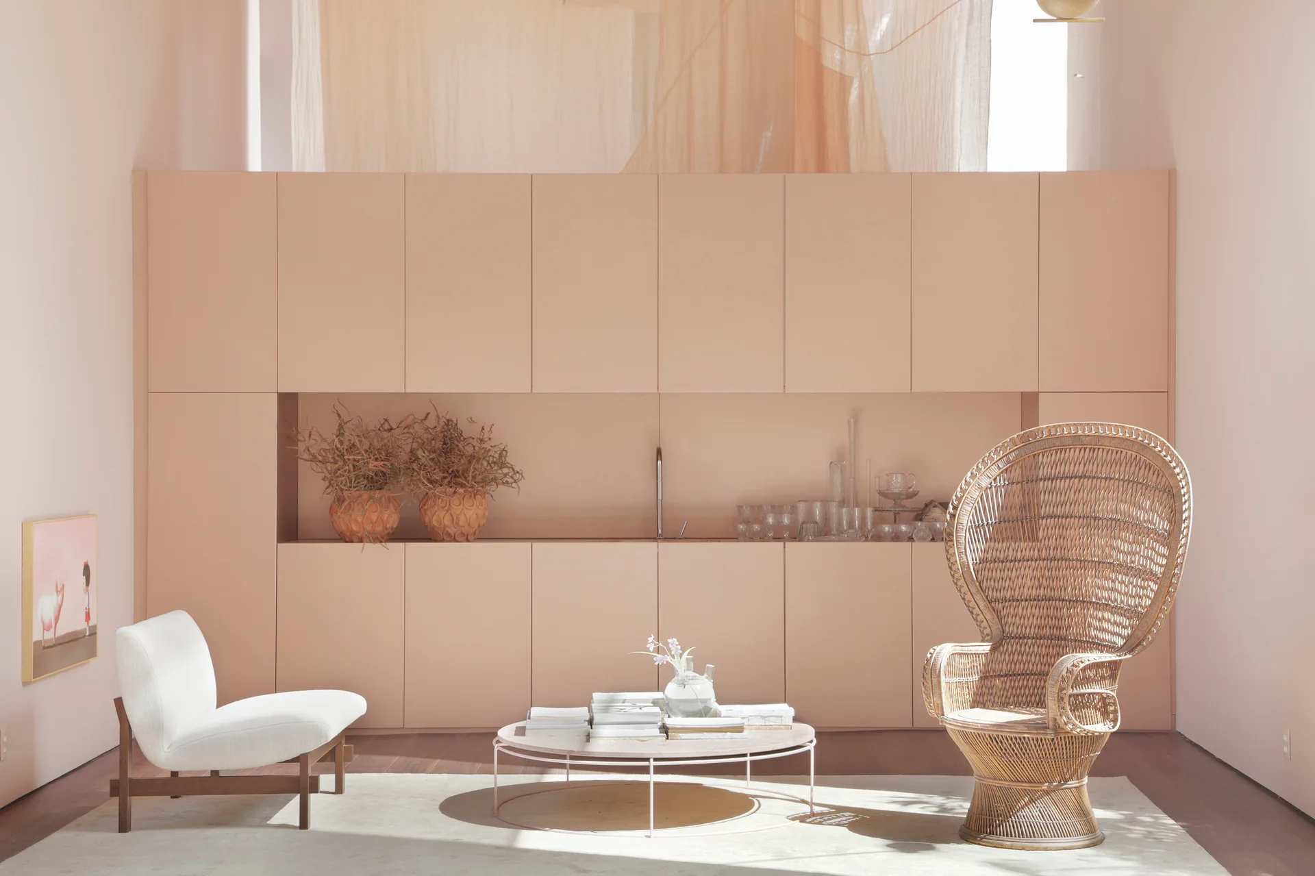 A minimalist living room features a full-wall blush pink cabinet system, a tall rattan peacock chair, and a low round coffee table on a light rug.