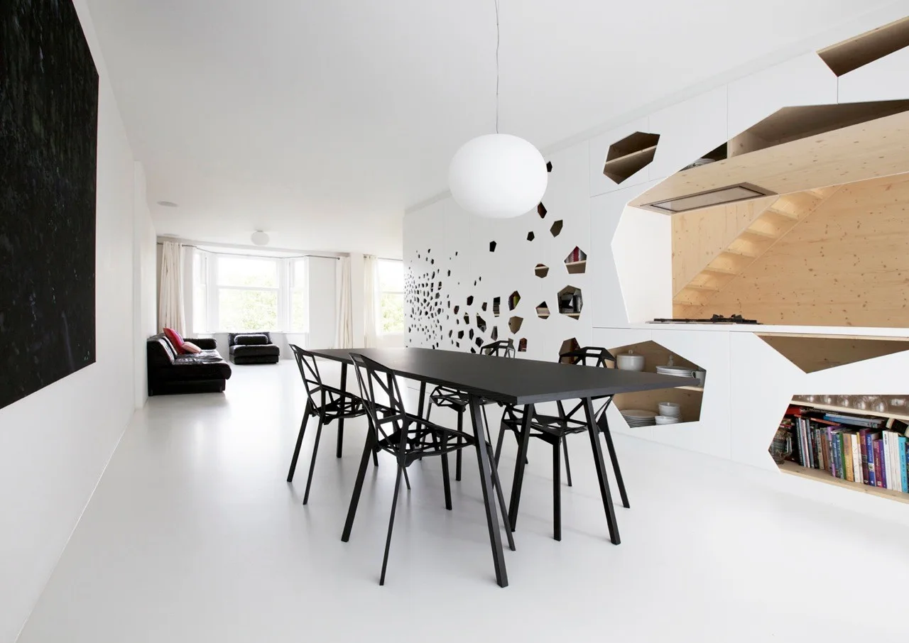 A black dining table and geometric chairs sit in a minimalist white room next to a storage wall with hexagonal cutouts revealing wood shelving.