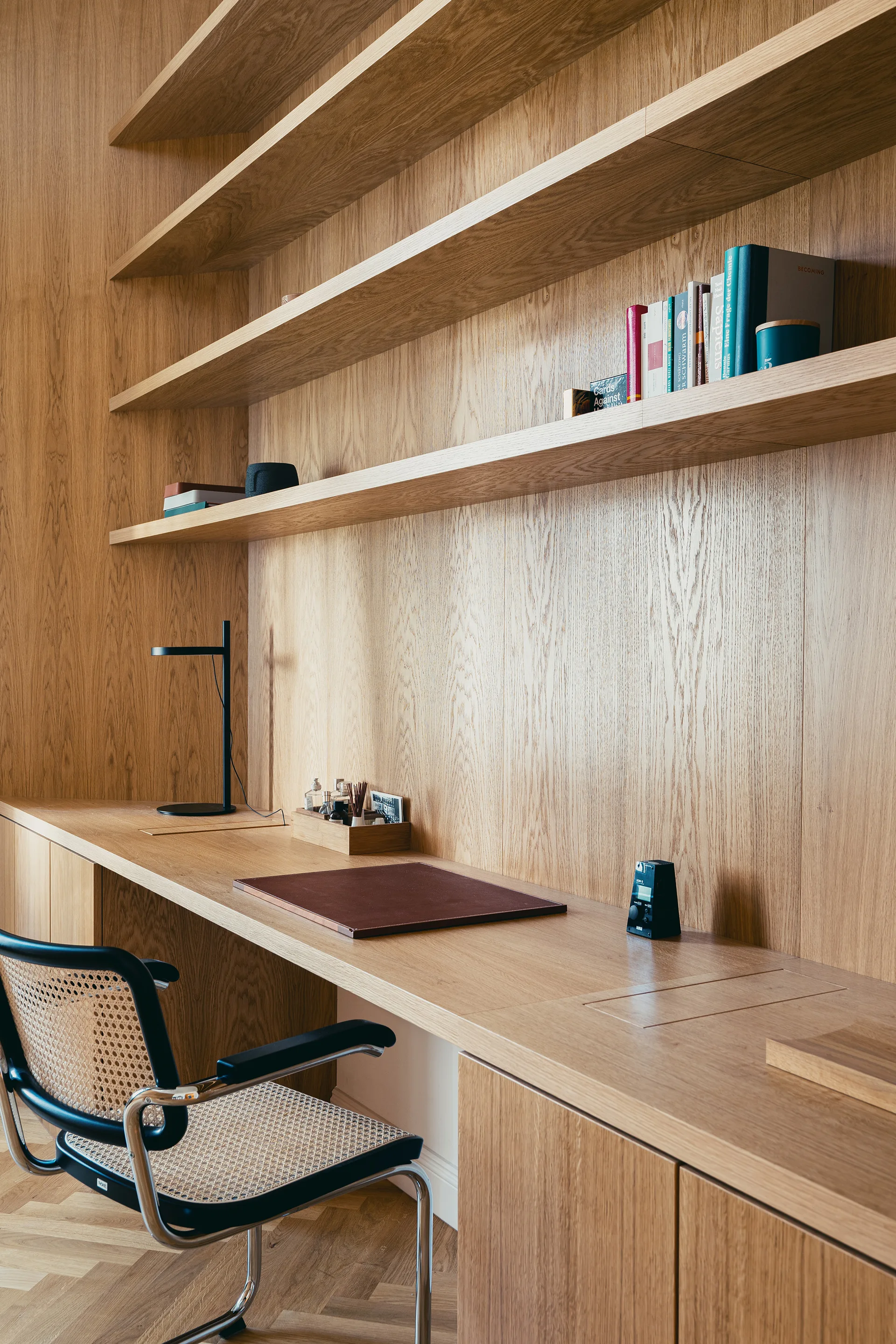 A home office features a long light oak built-in desk with matching wall paneling, open wood shelving, and a black cane chair on herringbone flooring.