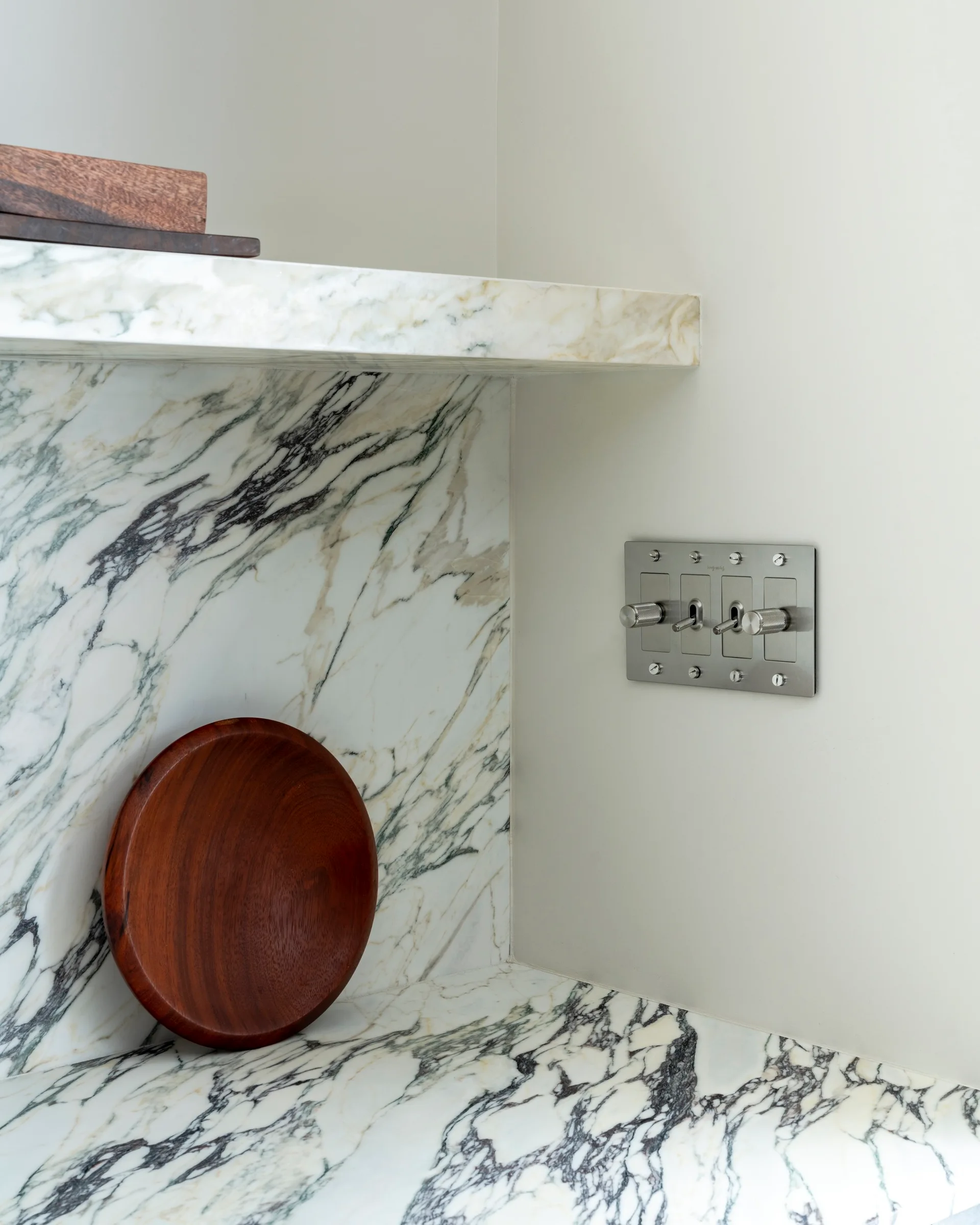 A kitchen corner features a veined white marble countertop and backsplash, a floating stone shelf, and a brushed metal toggle switch plate.