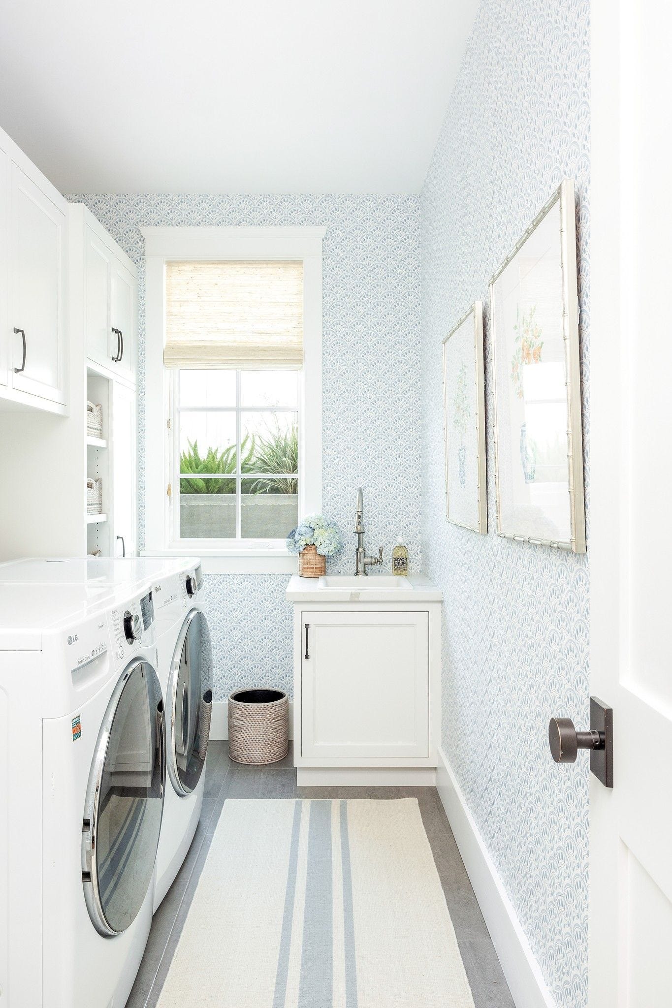 A bright laundry room featuring blue and white patterned wallpaper, white front-load washer and dryer, a utility sink, and a striped rug.
