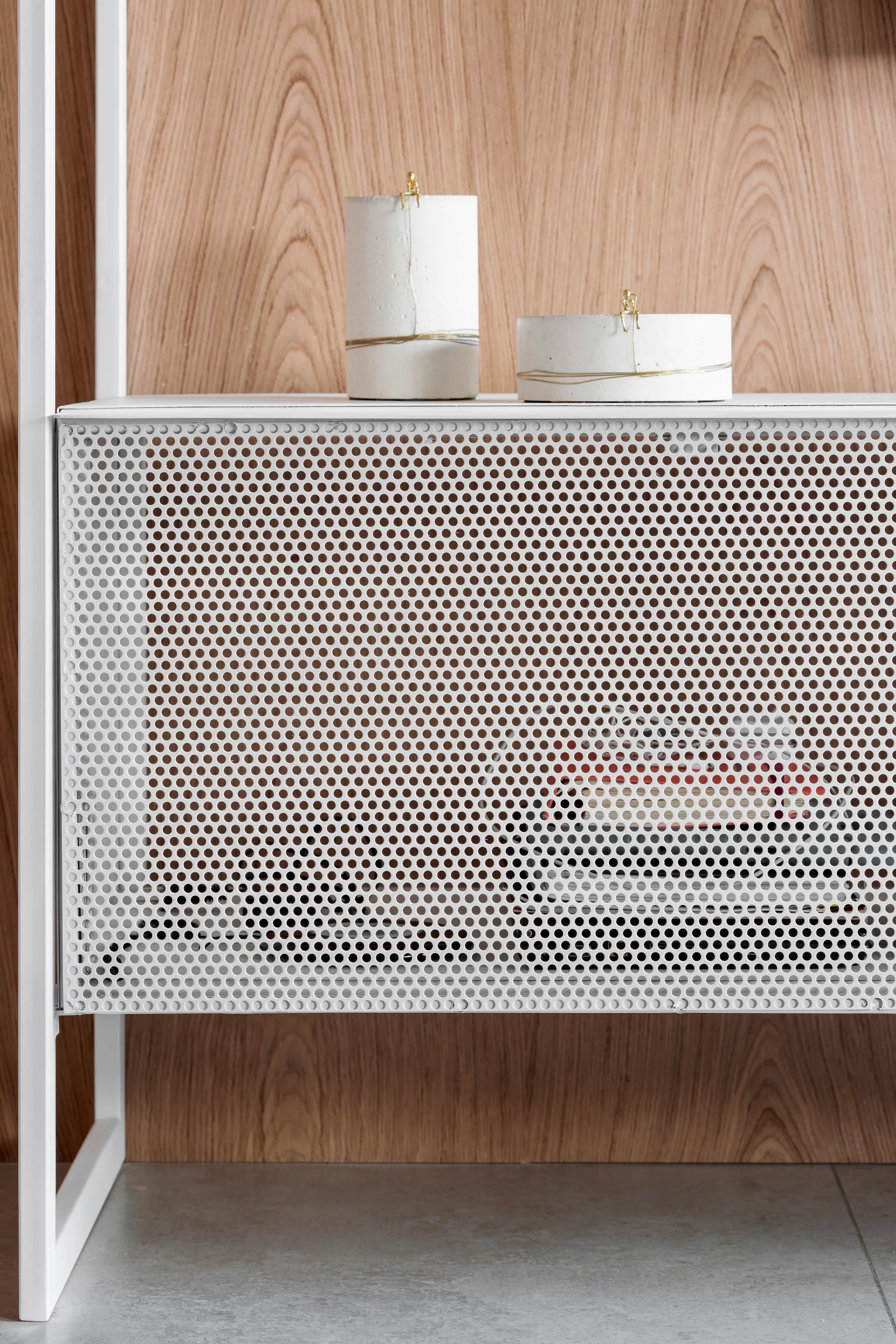 A white perforated metal cabinet with decorative cement canisters sits against a light wood paneled wall on a grey tiled floor.