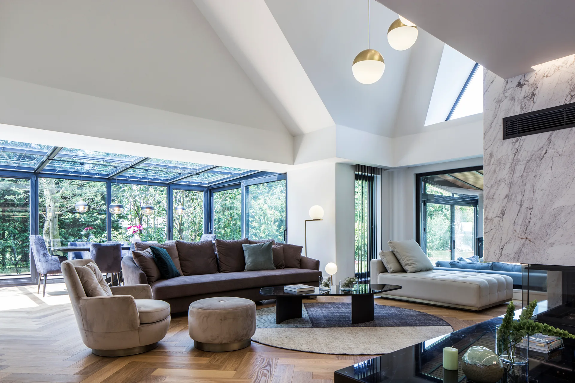 A spacious living room featuring a vaulted ceiling, herringbone wood floors, a brown sofa, and floor-to-ceiling glass walls looking out to a garden.