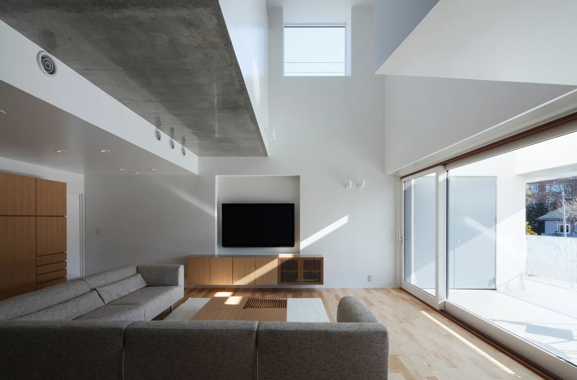 A minimalist living room features a grey sectional sofa, a floating light wood console, a wall-mounted TV, and an exposed concrete ceiling.