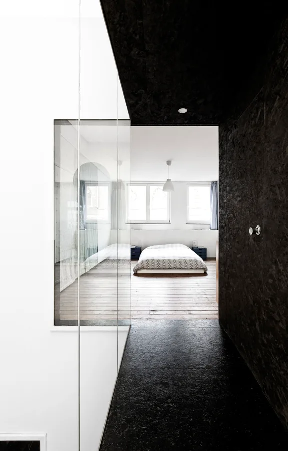 A view from a dark textured hallway through a frameless glass wall into a white bedroom with a low bed and wood flooring.