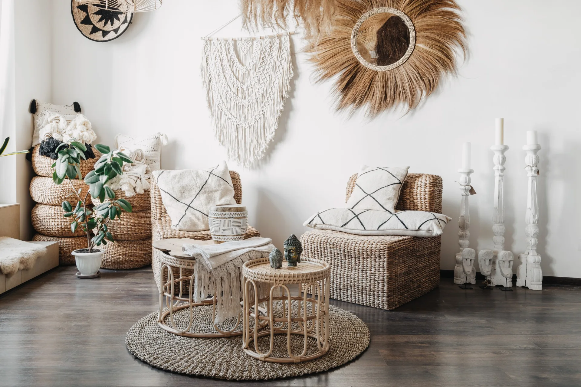 Two woven wicker armchairs stand on a dark wood floor near round wicker coffee tables, a circular jute rug, and a macrame wall hanging.