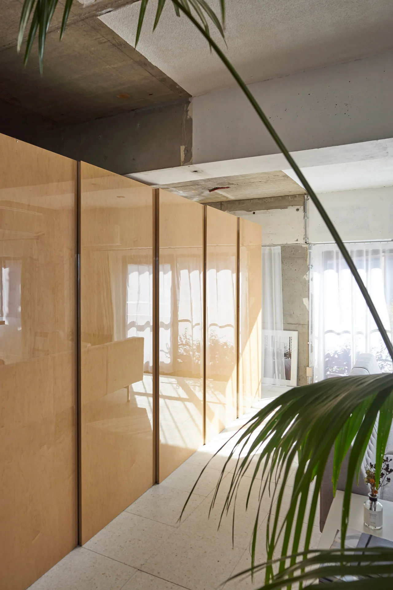 Large glossy wood-toned wardrobe cabinets stand against a raw concrete ceiling and light tile flooring near a window with sheer curtains.