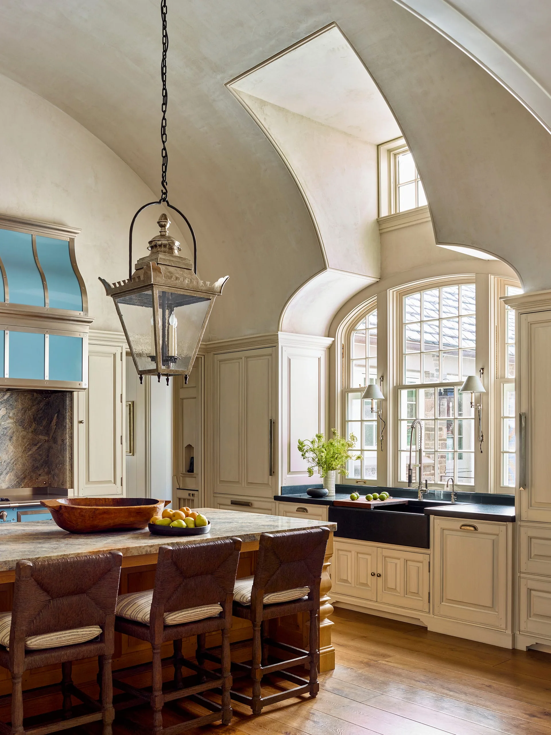 A French country kitchen features an arched plaster ceiling, cream cabinetry, a bright blue range hood, and a central stone island with woven stools.