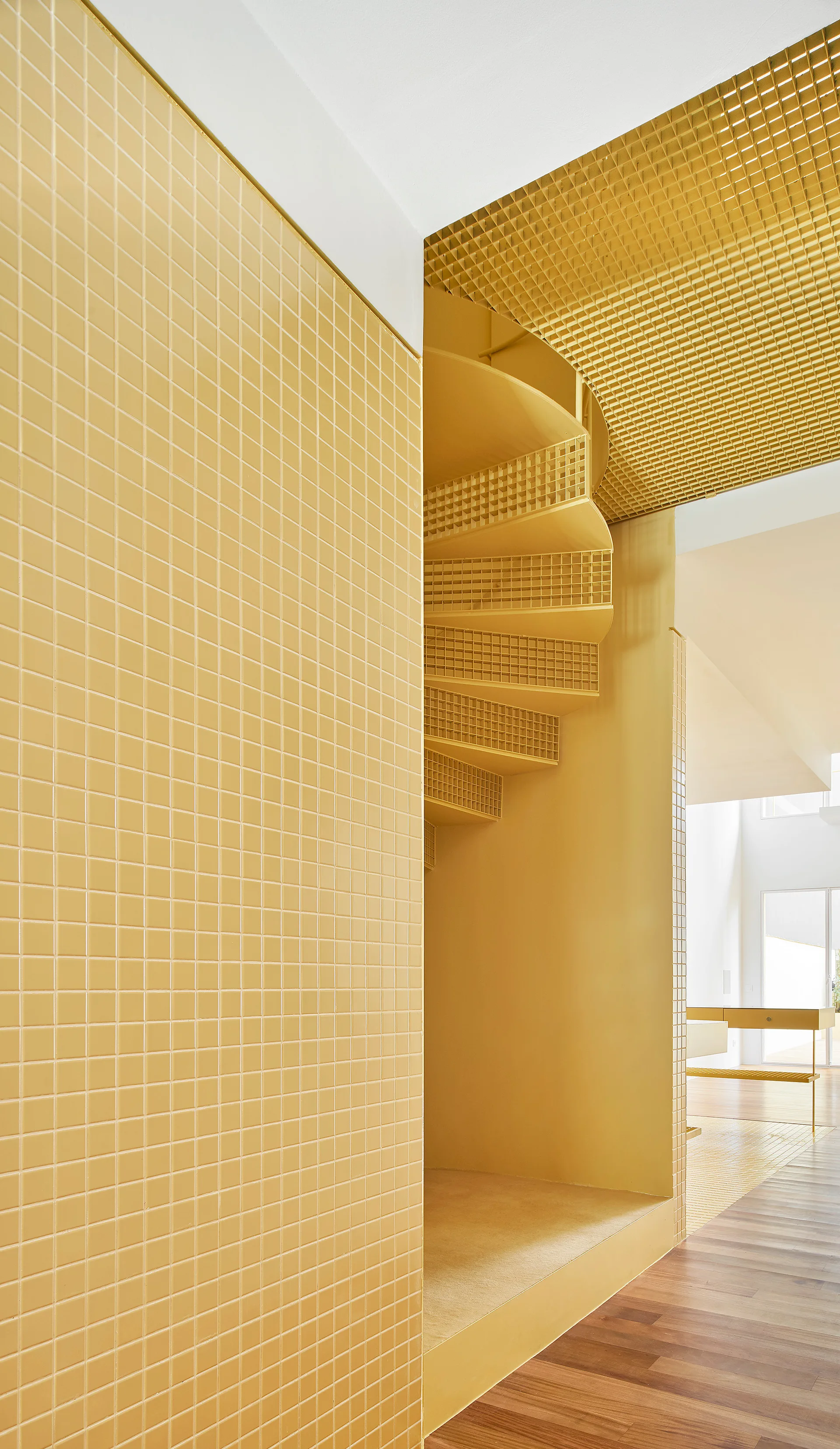 A yellow spiral staircase with grid steps stands next to a matching yellow tiled wall on a hardwood floor.