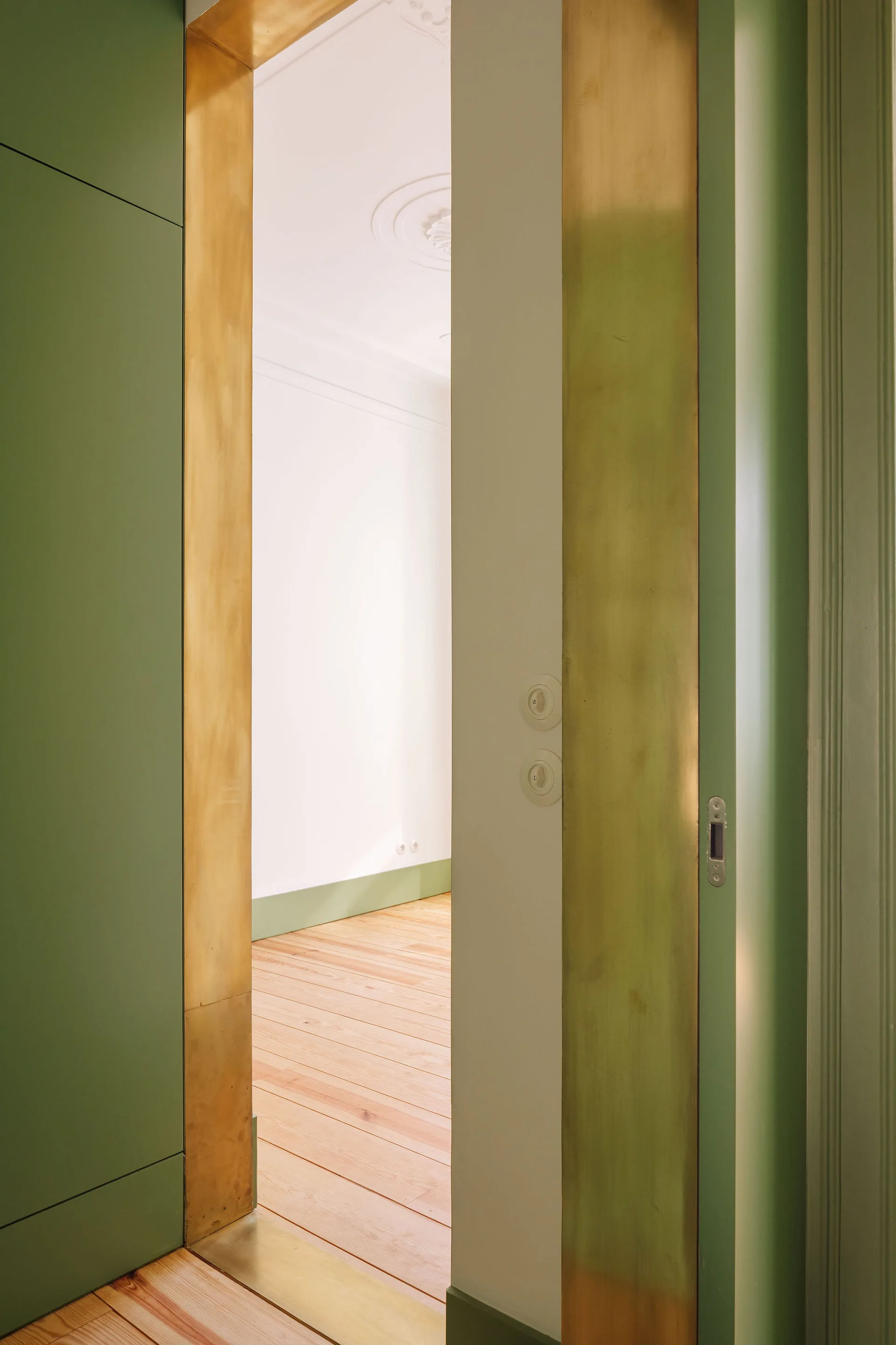 A close-up view through a brass-trimmed doorway surrounded by green wall panels looking into a bright room with light wood floors.
