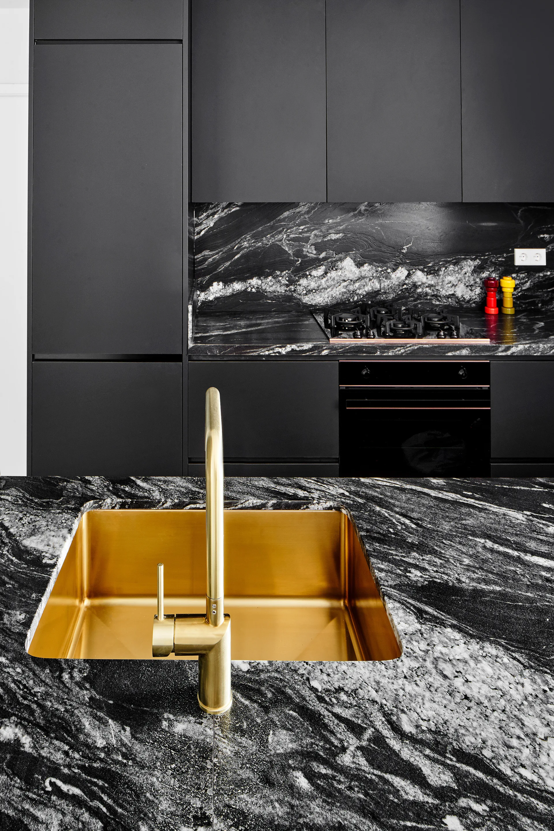 A modern kitchen details a veined black marble countertop and backsplash, matte black cabinets, and an integrated gold undermount sink and faucet.