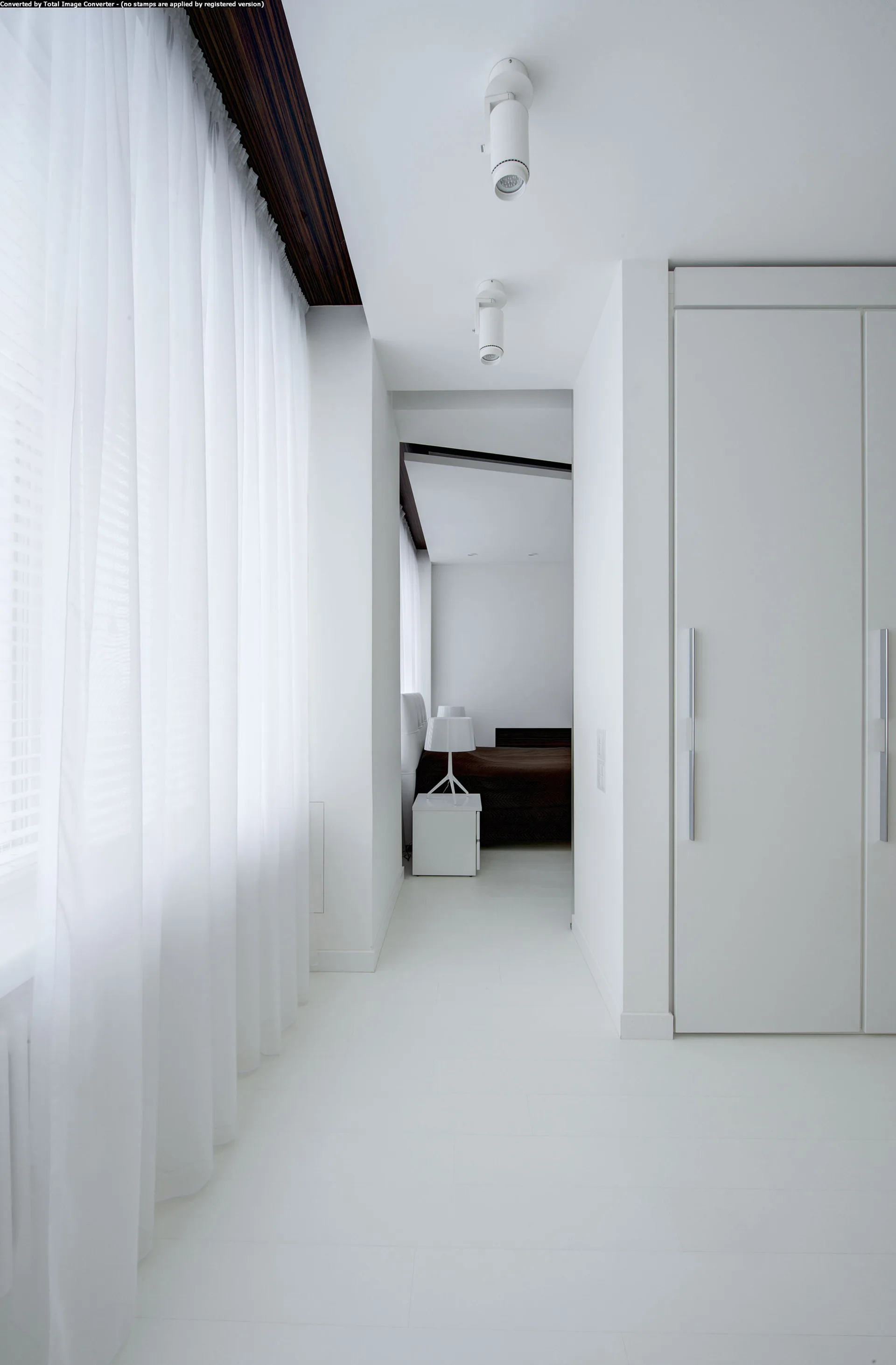 A minimalist white hallway with sheer floor-to-ceiling curtains, a large built-in wardrobe, and ceiling spotlights leading into a bedroom.