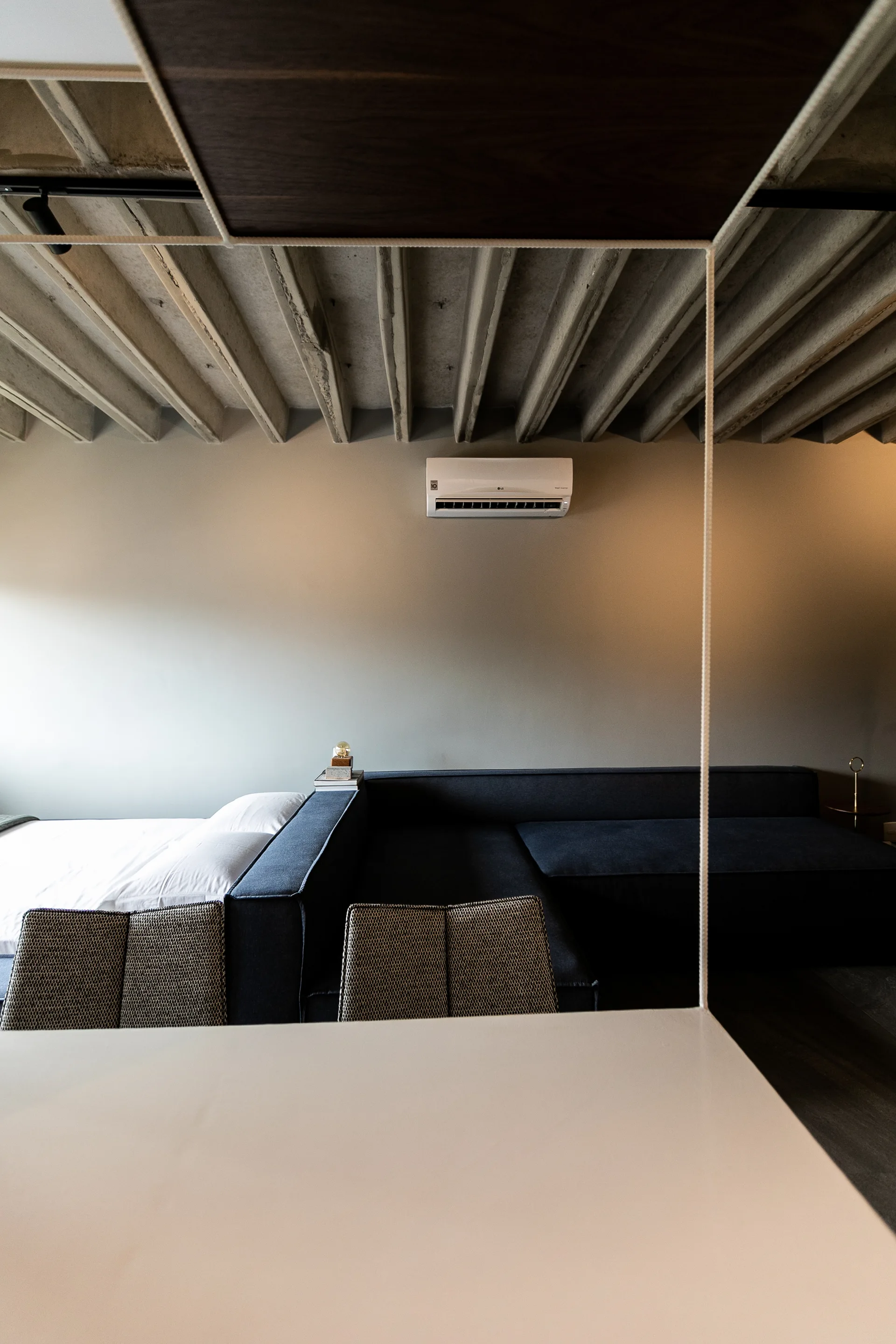 A modern bedroom features an exposed concrete ceiling with ribbed joists, a dark modular sofa, and a bed with white bedding.