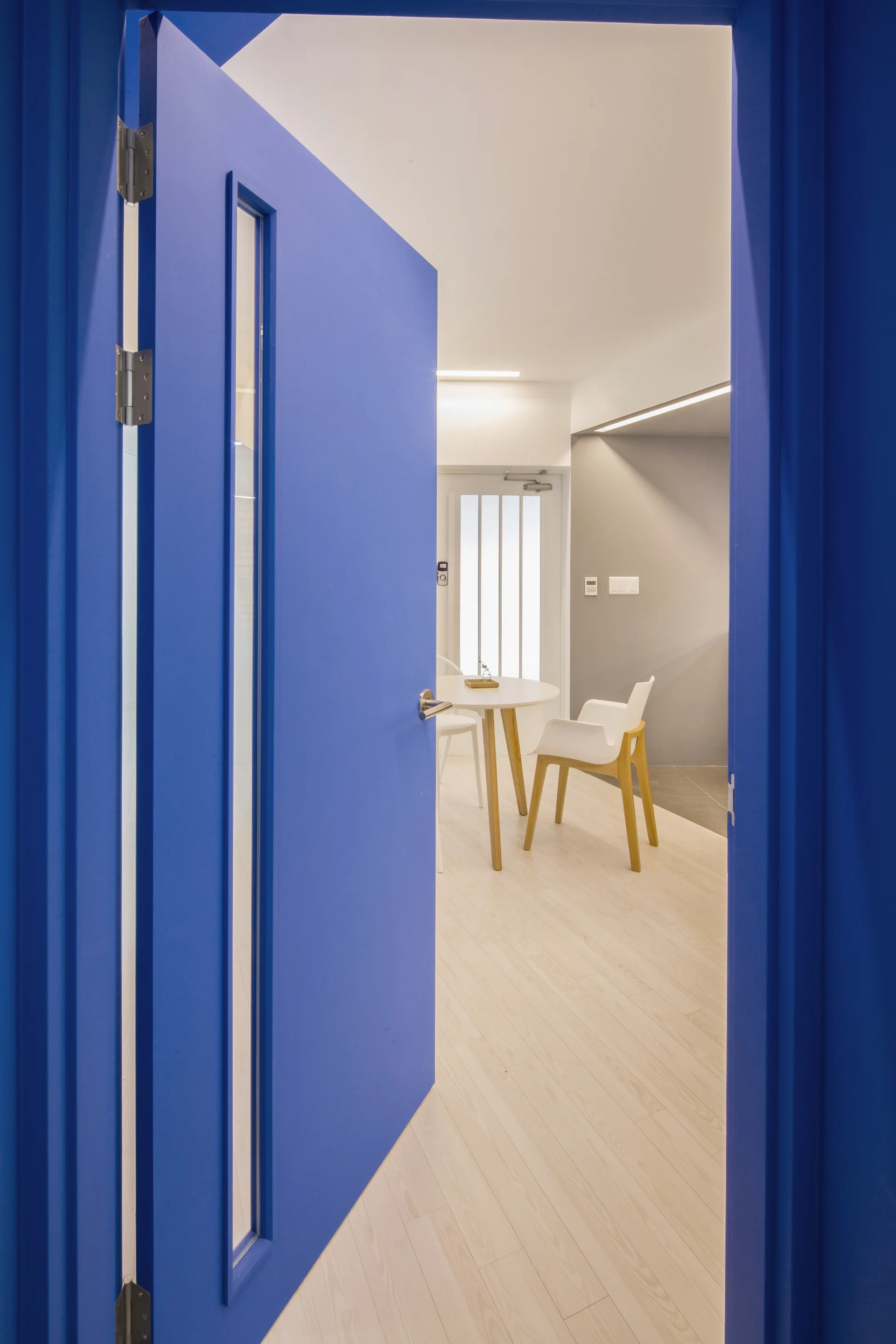 A bright blue interior door with a vertical glass pane opens to reveal a minimalist dining room with a white table and chairs.