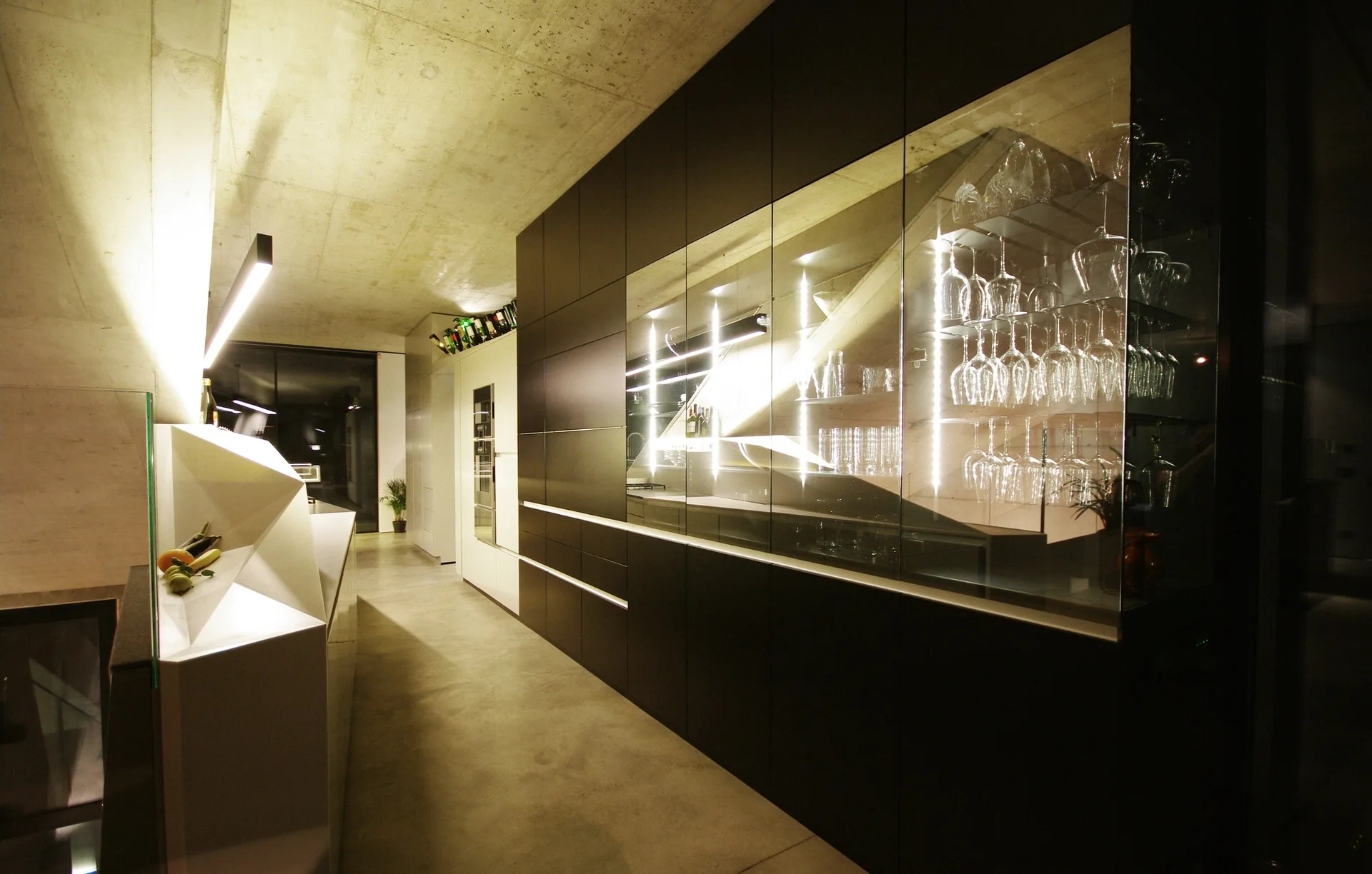 A modern kitchen featuring a sculptural white island, concrete floors, and a matte black cabinet wall with an integrated illuminated glass display case.