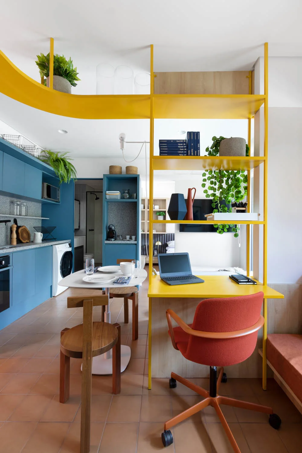 A small kitchen with blue cabinets stands beside a workspace containing a yellow metal shelving unit, an orange office chair, and terracotta floor tiles.