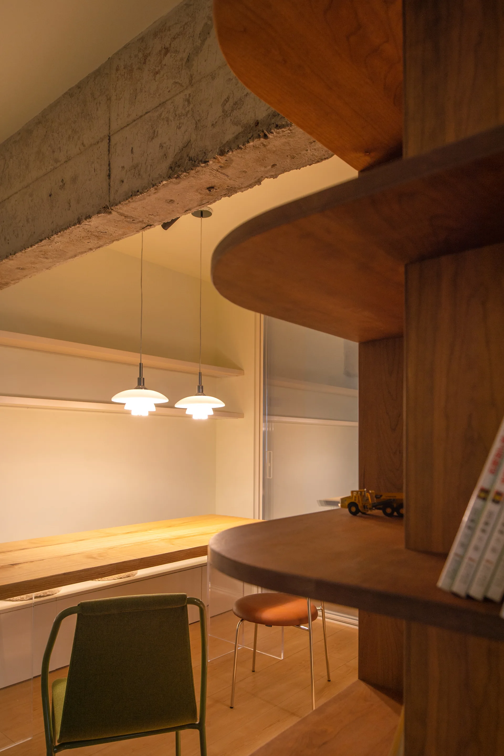 A minimalist workspace featuring a long wood desk, a green chair, two hanging pendant lights, and a prominent exposed concrete beam overhead.