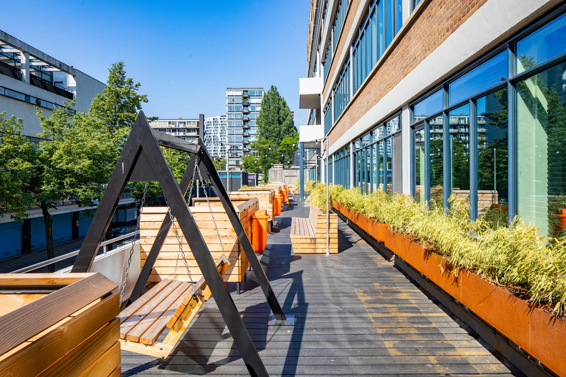 A long outdoor wooden deck terrace features wooden swing seats, matching benches, and rust-colored metal planters filled with ornamental grasses.