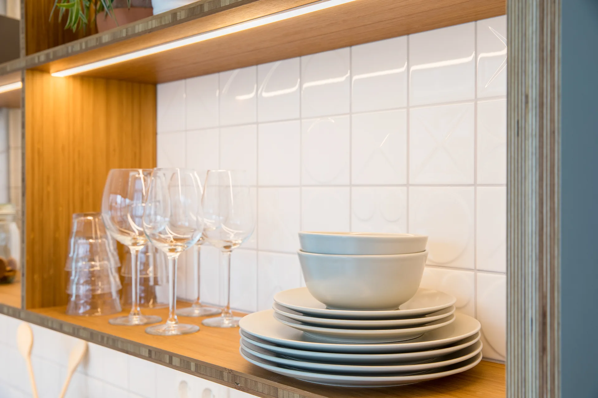 Open wooden kitchen shelving holds stacked white dinnerware and stemware against an embossed white tile backsplash illuminated by warm LED lighting.