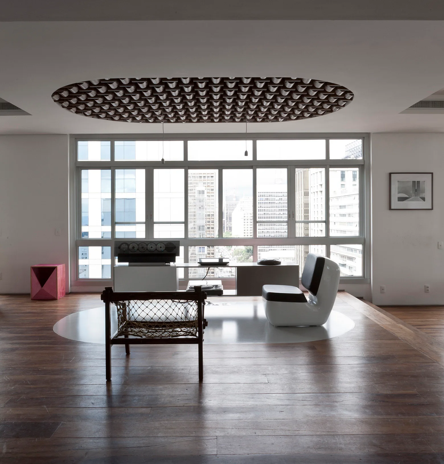 A minimalist living room with hardwood floors, a textured oval ceiling installation, and modern seating facing large windows with a cityscape view.