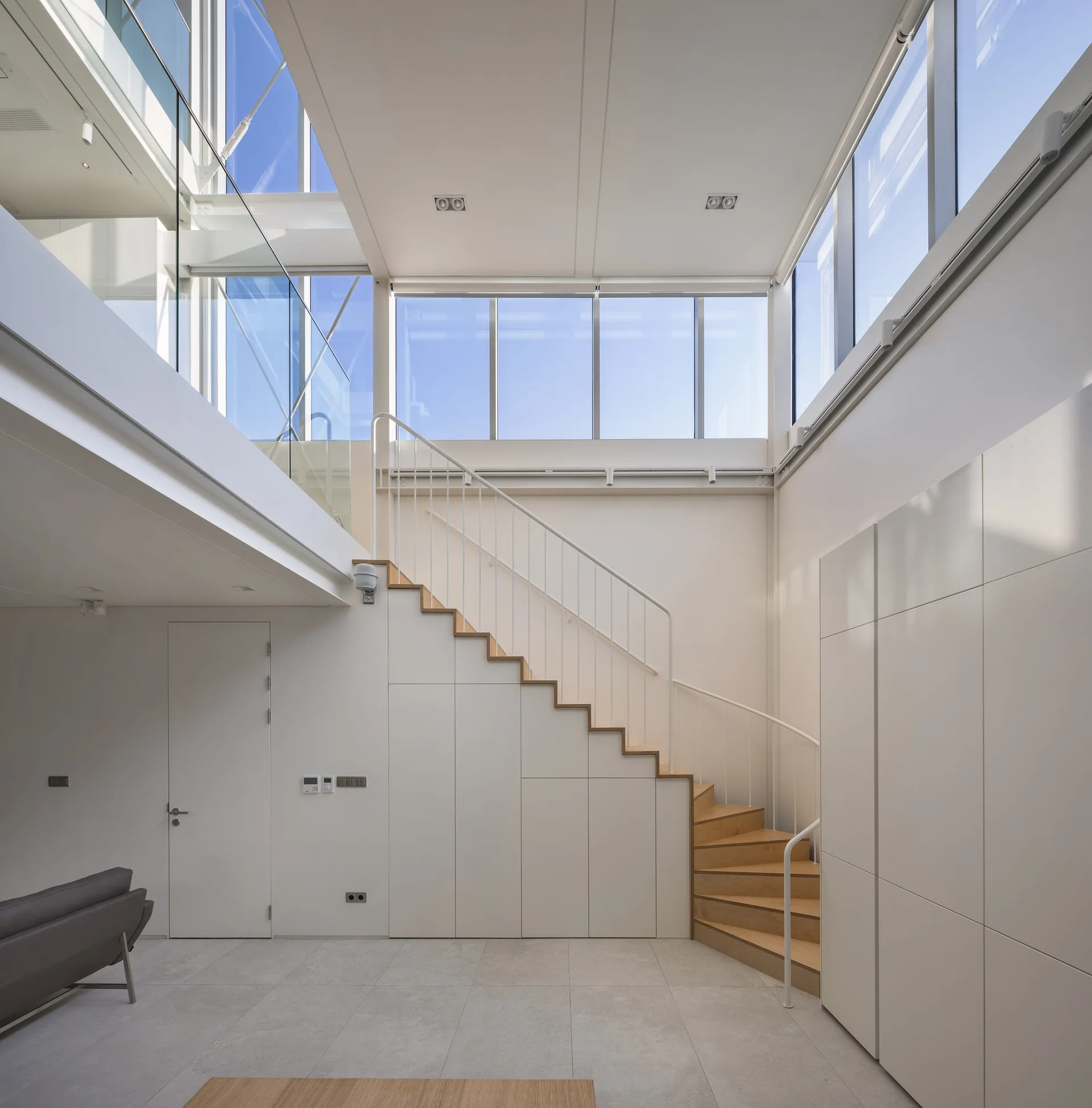 A minimalist double-height room features a light wood staircase with integrated white cabinets underneath, a metal railing, and high windows.