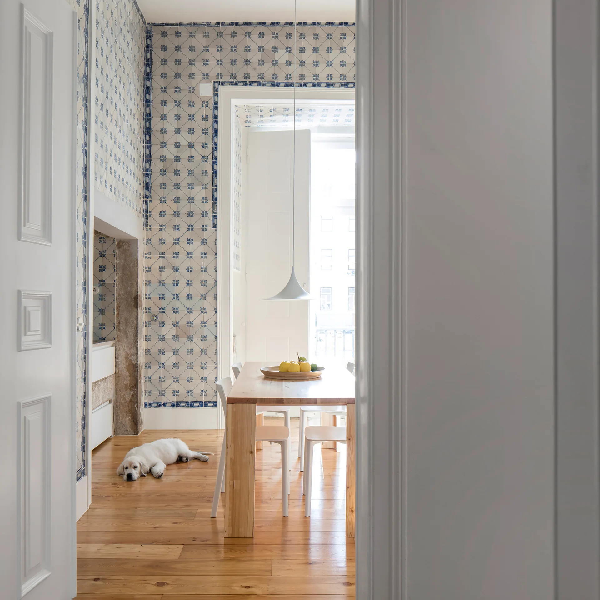 A dining room seen through a white doorway featuring a light wood table with white chairs and walls covered in blue and white patterned tiles.