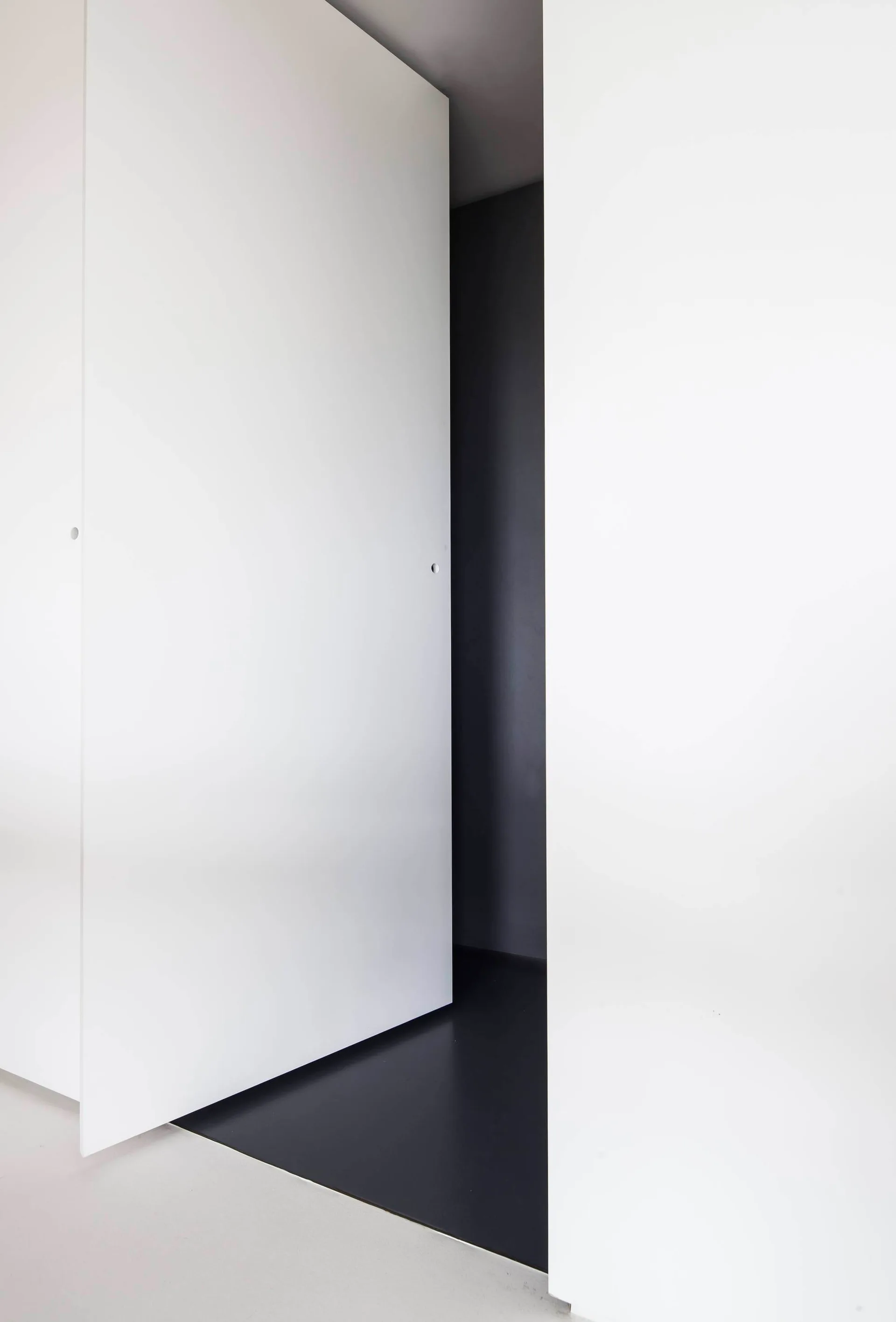 A matte white floor-to-ceiling pivot door slightly ajar, opening into a dark gray interior hallway with dark floors.