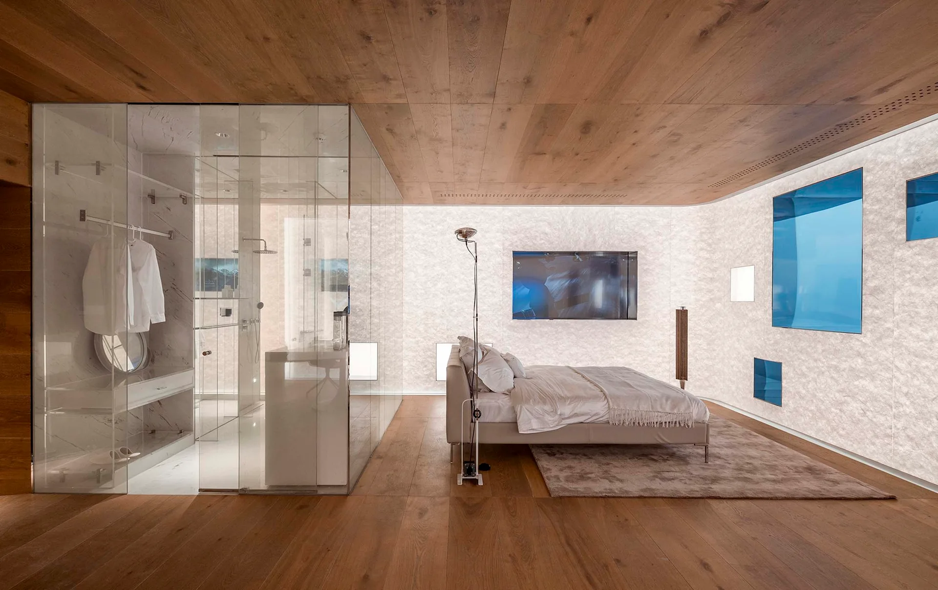 A minimalist bedroom featuring a low-profile bed on a rug, a wood plank ceiling and floor, and a glass-enclosed bathroom partition on the left.