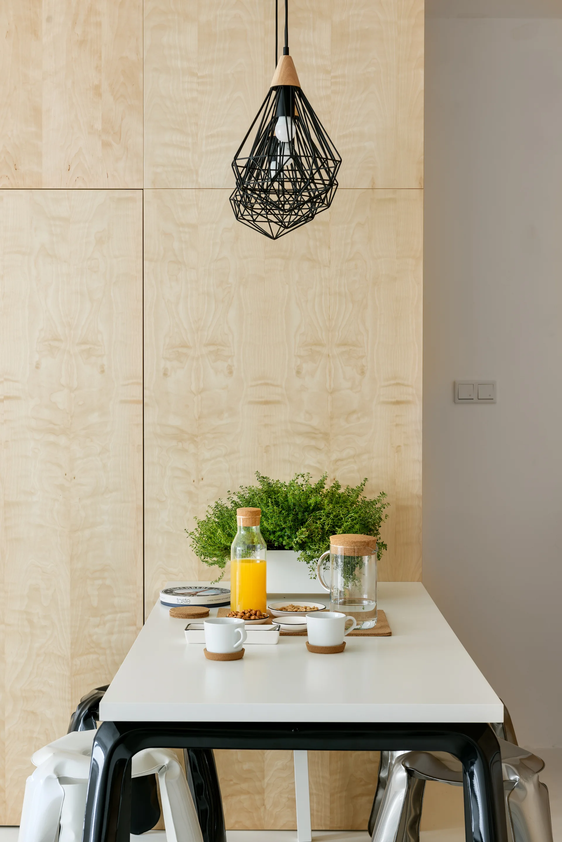 A white dining table with drinks and mugs sits against a light wood-paneled wall under a black geometric pendant light.