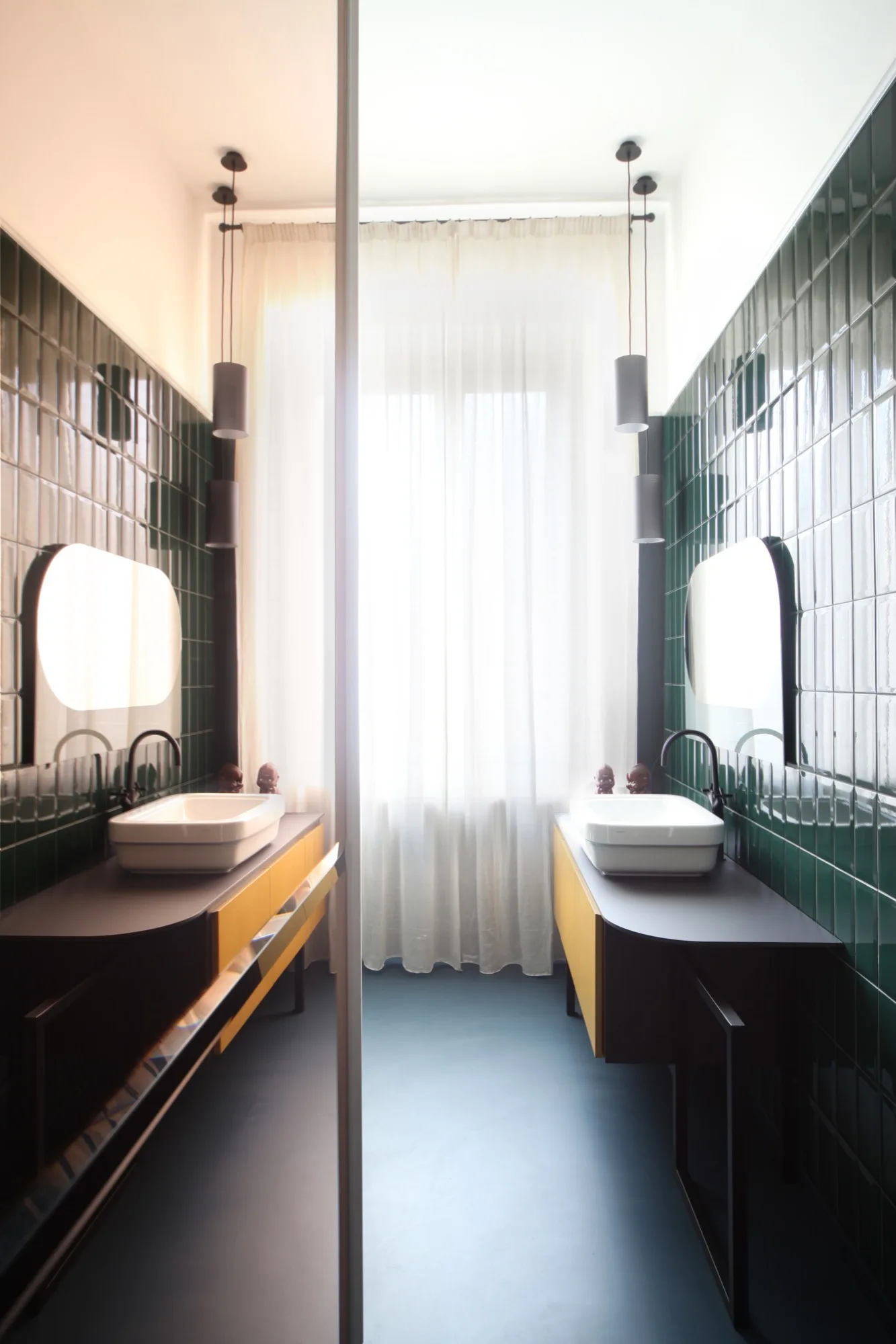 Symmetrical bathroom with two yellow vanities with white vessel sinks set against glossy deep green tiled walls flanking a sheer curtained window.