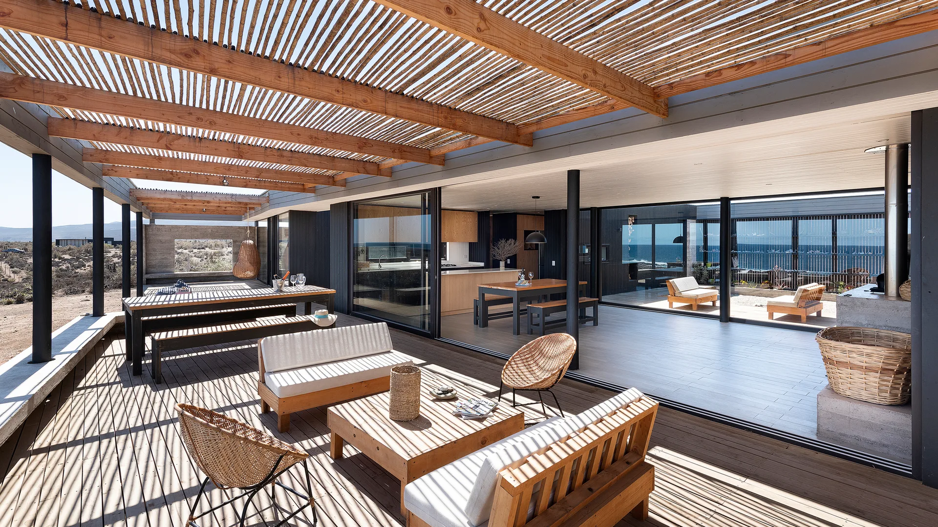 A wooden deck with a slatted pergola overhead features wood outdoor furniture and large sliding glass doors leading into the house.