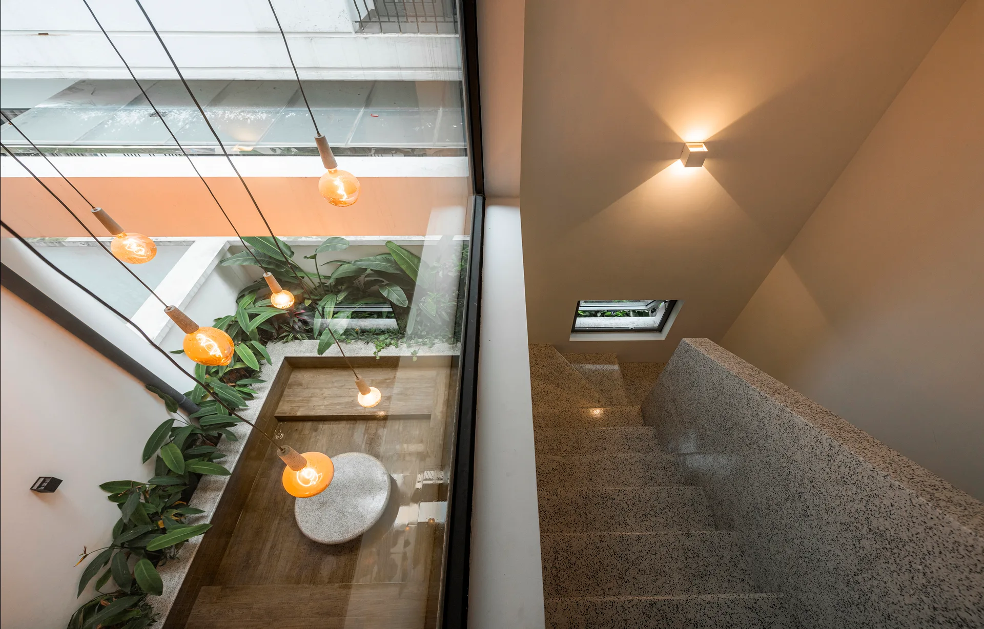 A high-angle view of a speckled terrazzo staircase beside a large window looking into a vertical atrium with hanging warm pendant lights and plants.