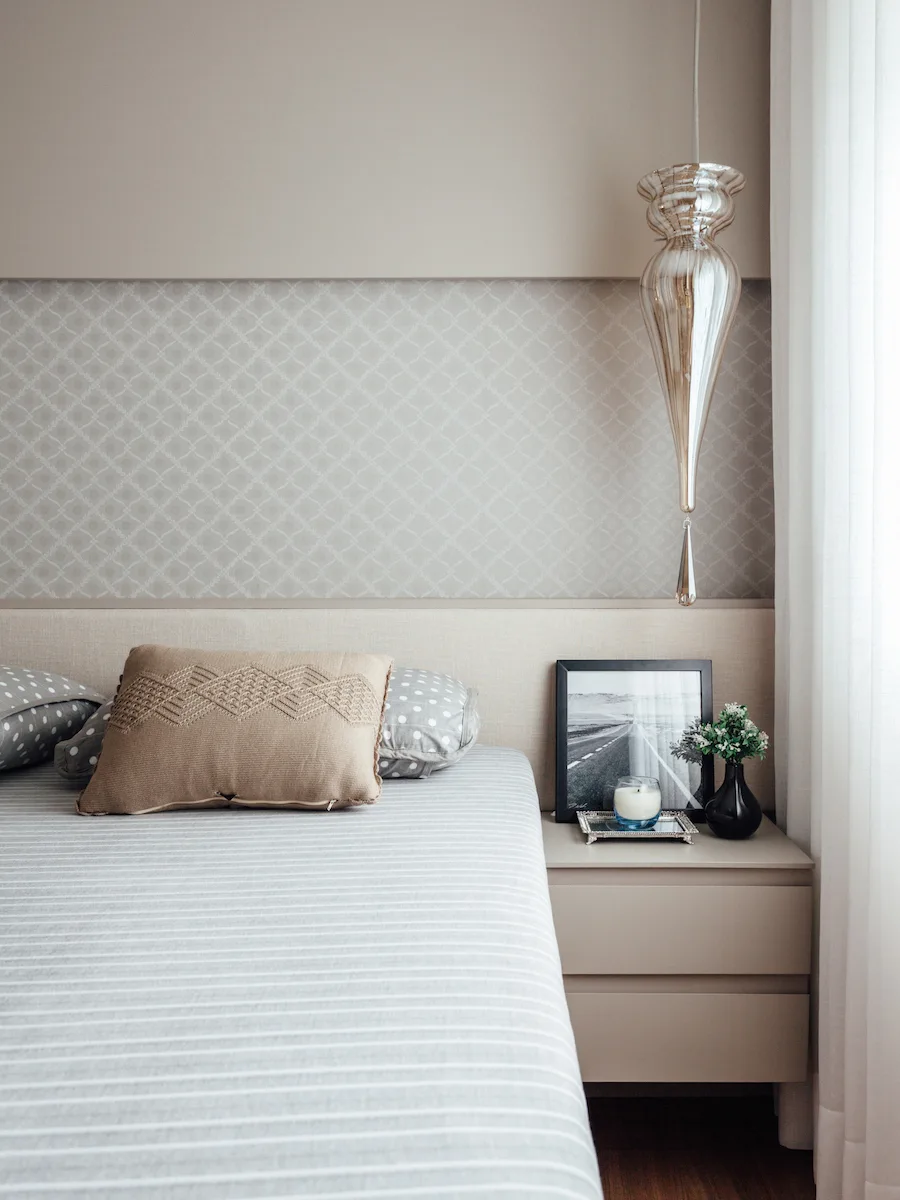 A modern bedroom features a bed with striped bedding, a taupe nightstand, patterned wallpaper, and a hanging glass pendant light.