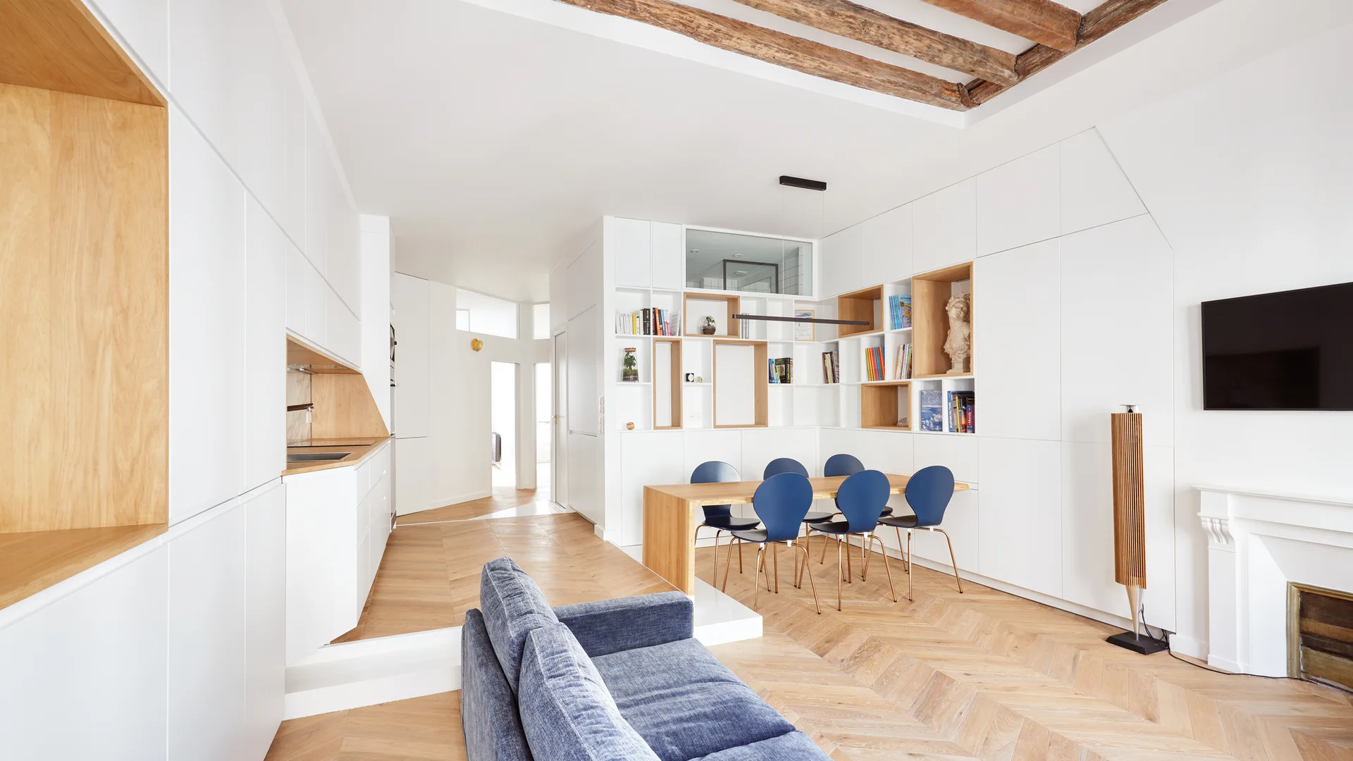 An open-plan living and dining area featuring white built-in storage cabinets with wood niches, a wooden dining table, blue chairs, and chevron floors.
