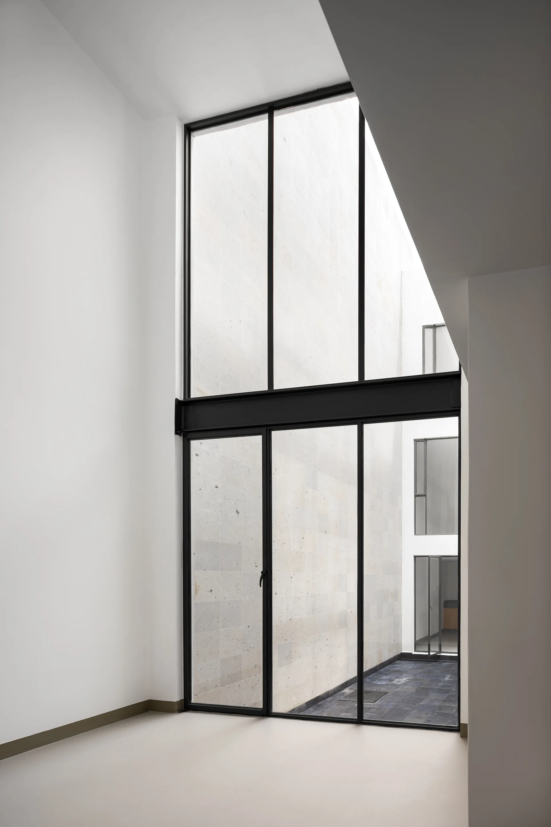A towering double-height black metal framed window in a minimalist white room looks out onto a stone-walled courtyard.