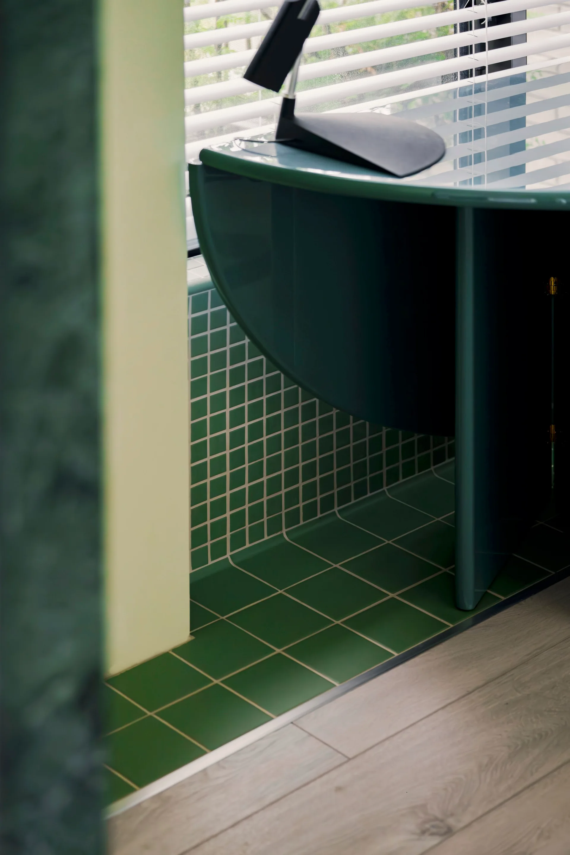 A curved teal desk stands next to a green tiled recess by a window with white horizontal blinds.
