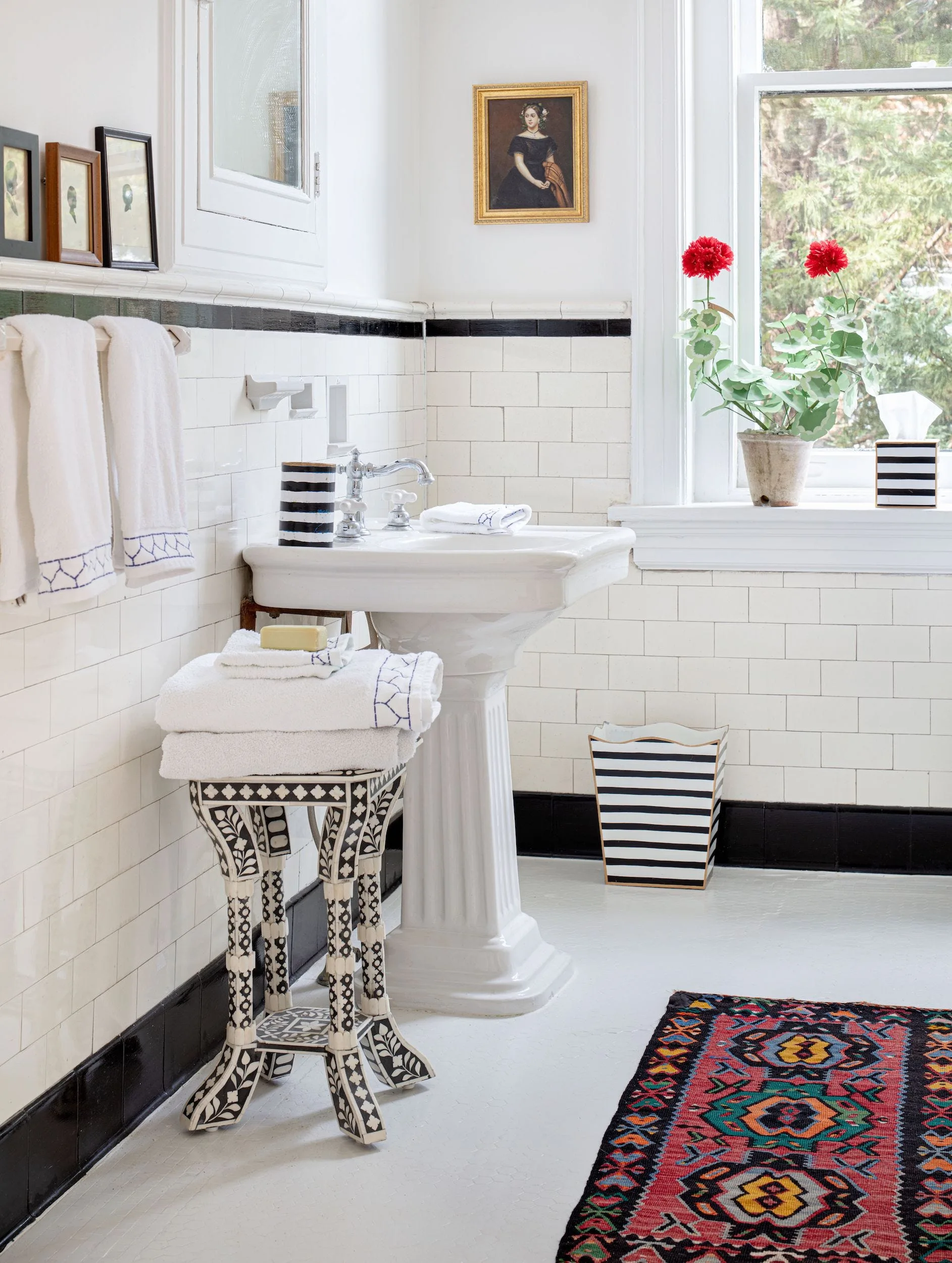 A white pedestal sink stands against subway tiled walls with black trim, flanked by a patterned inlay stool and a colorful patterned rug.