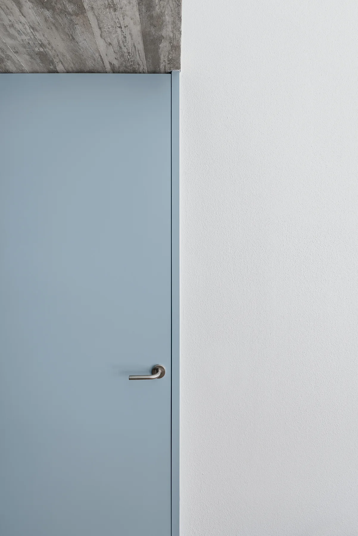 A minimalist light blue flush door with a brushed metal handle sits beneath a raw concrete ceiling next to a white wall.