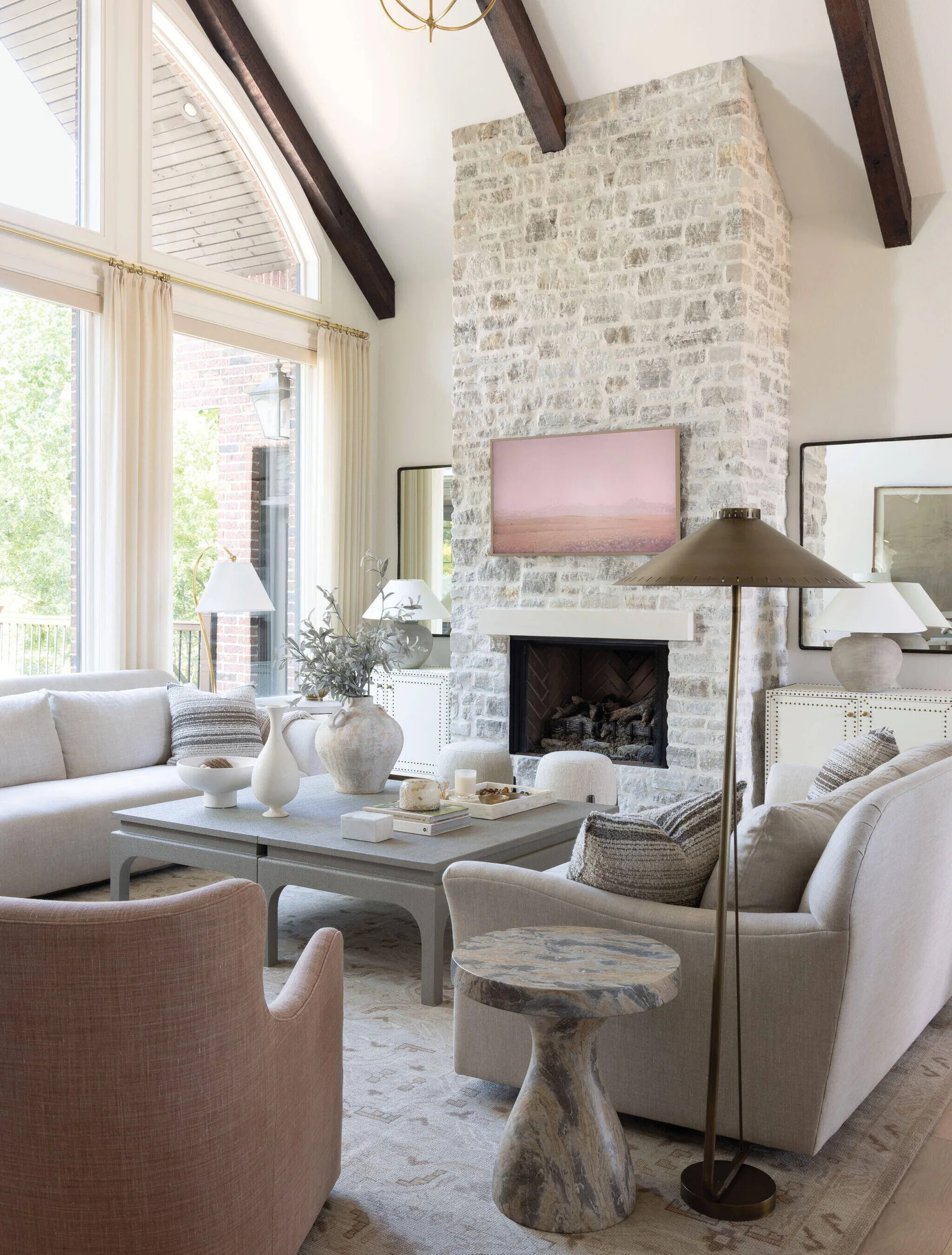 A bright transitional living room featuring a tall white stone fireplace, dark wood ceiling beams, and symmetrical sofas around a grey coffee table.