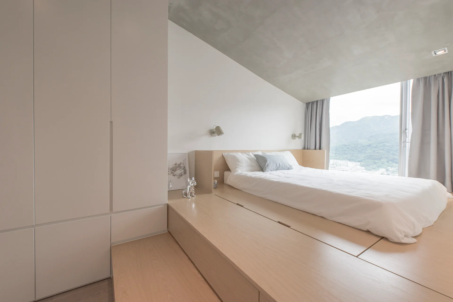A minimalist bedroom featuring a bed on a raised light wood platform next to white built-in wardrobes under an angled concrete ceiling.