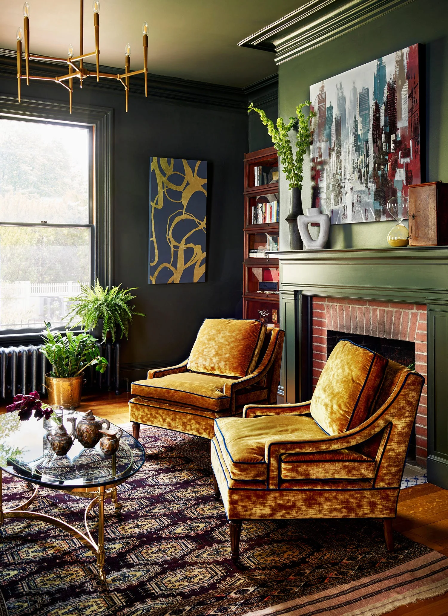 A green living room features two gold velvet armchairs, a brick fireplace with a matching green mantel, a patterned rug, and a glass coffee table.