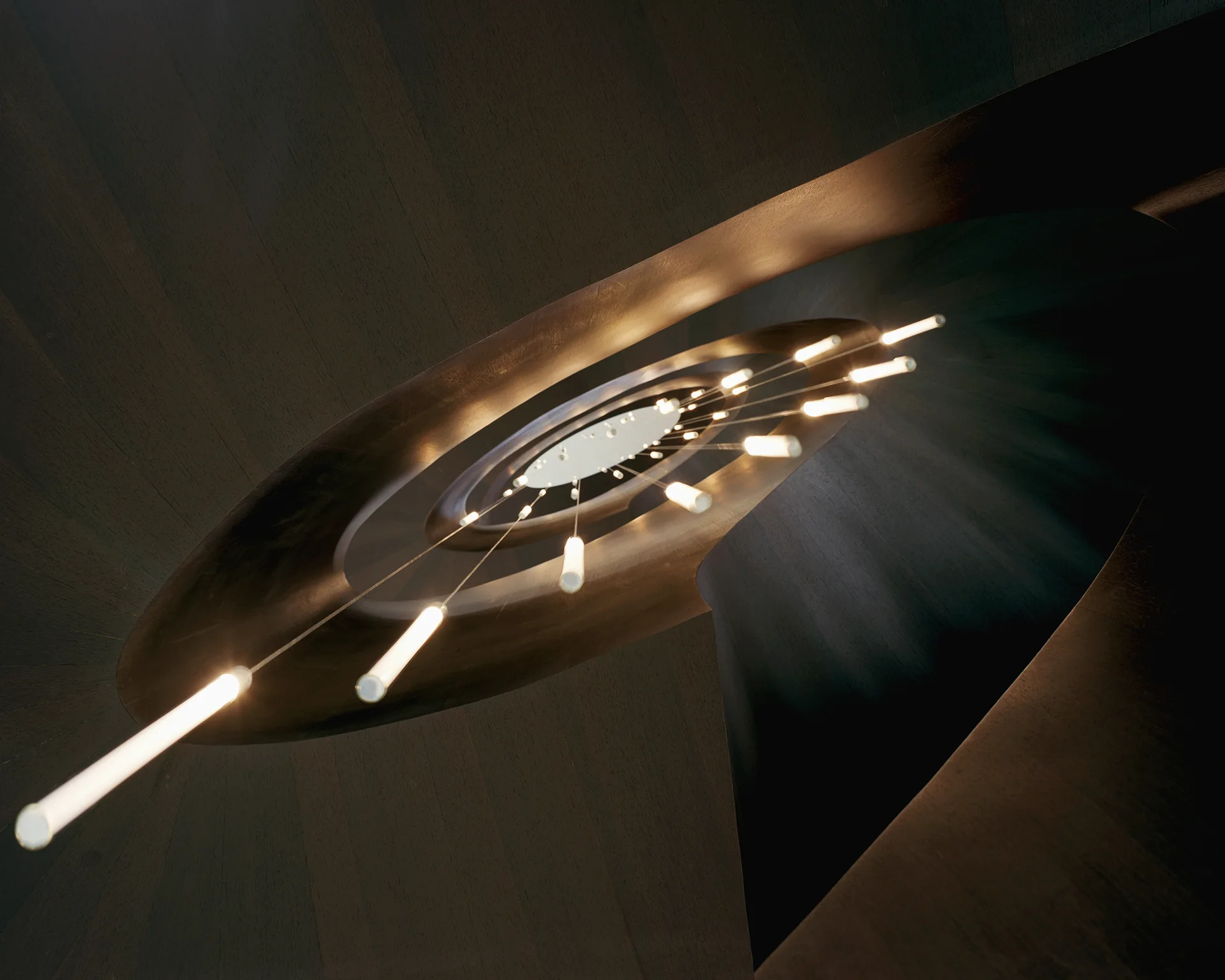 A low-angle view looking up a dark brown wood spiral staircase featuring slender cylindrical pendant lights suspended at various heights.