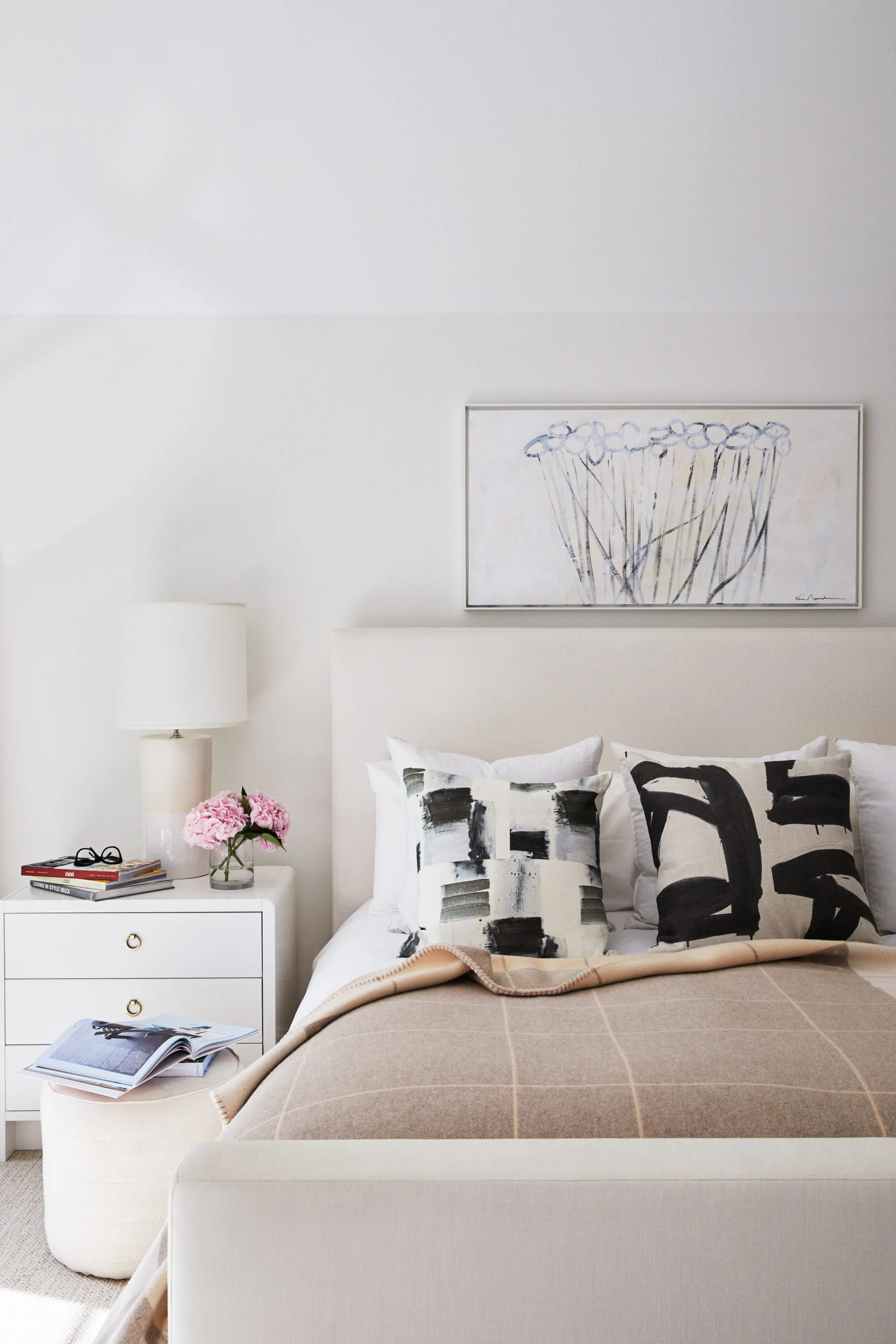 A cream bedroom features an upholstered bed with abstract black and white pillows, a white three-drawer nightstand, and a ceramic table lamp.