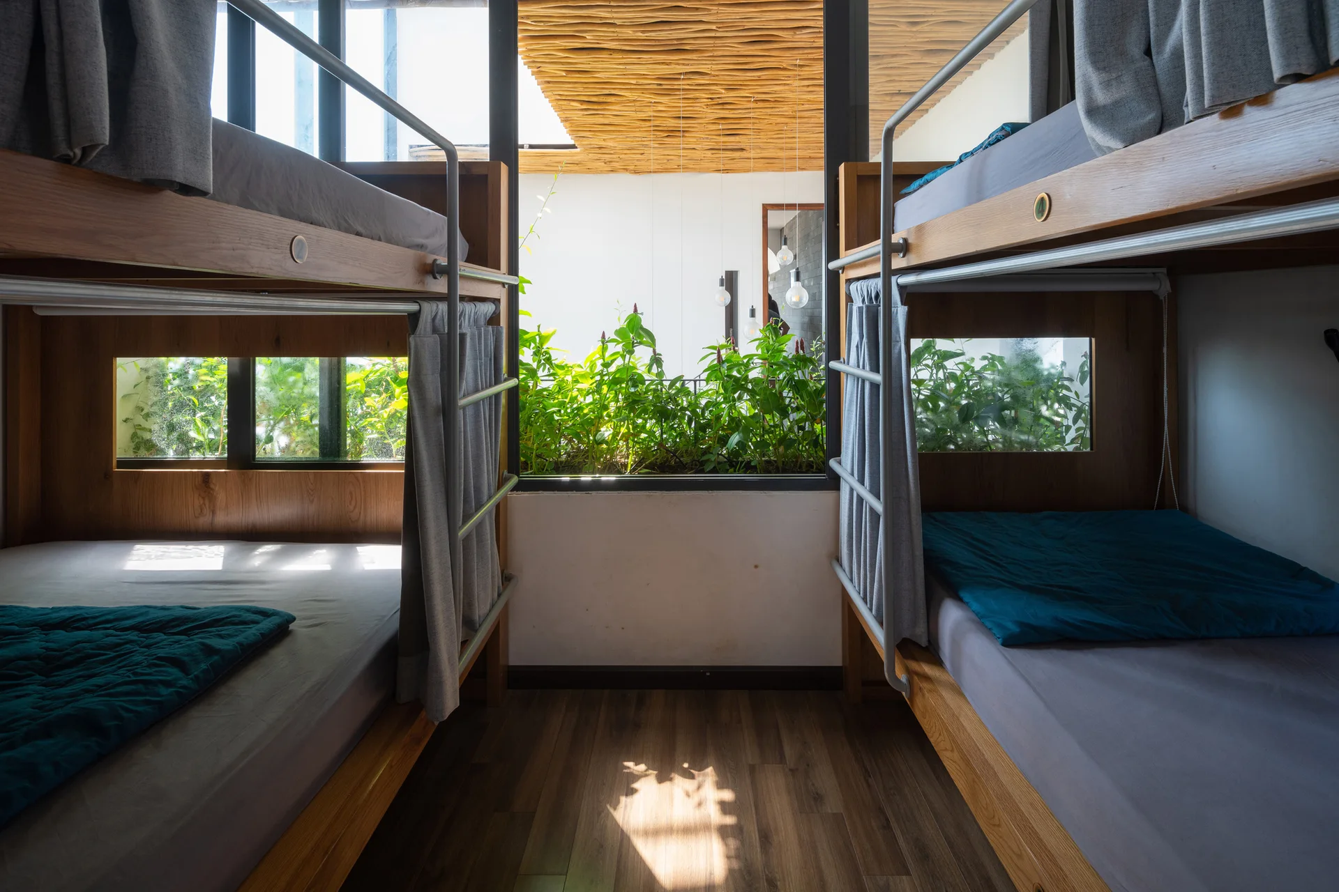 A shared bedroom featuring two wooden bunk beds with grey curtains facing a central window overlooking lush green indoor plants.