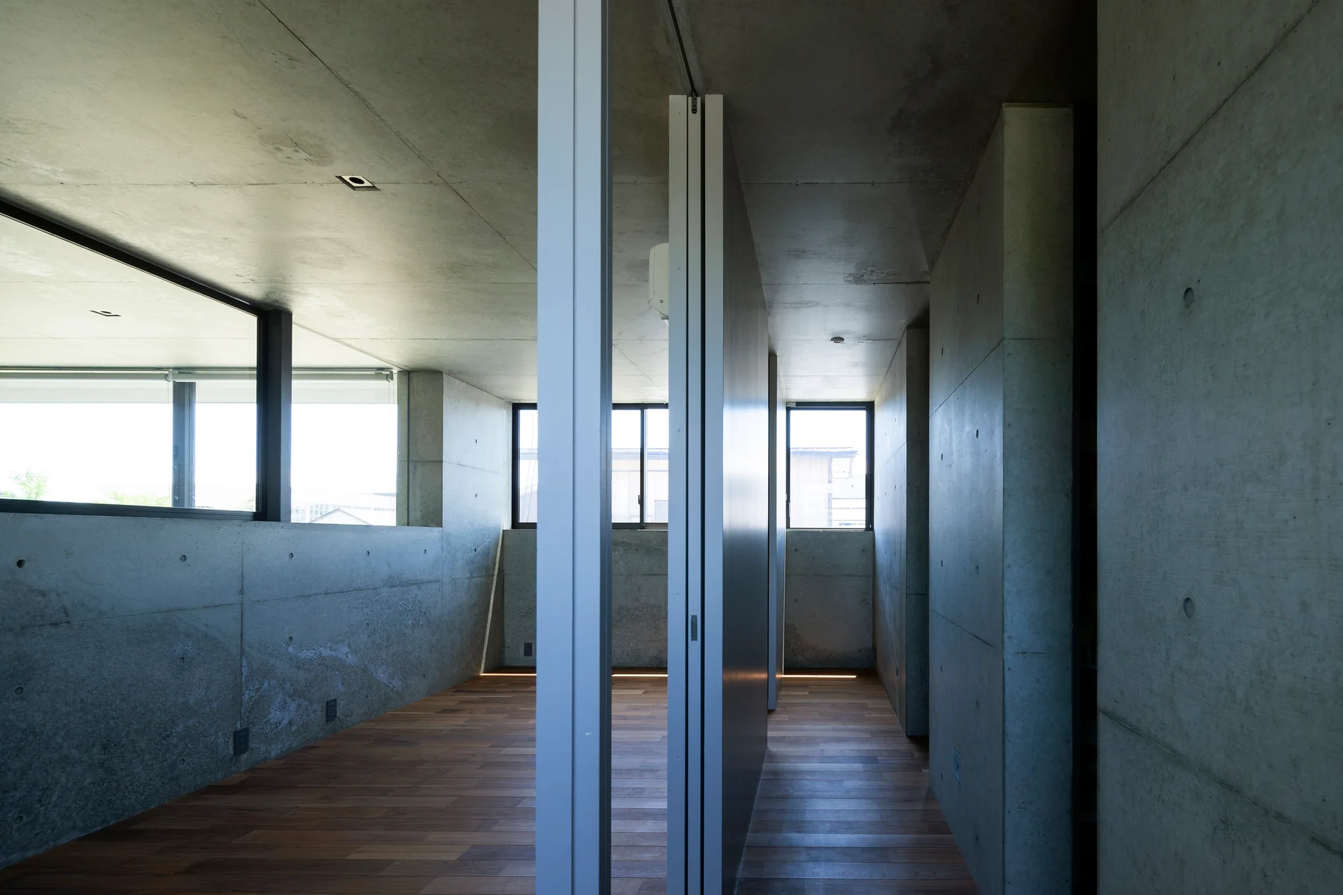 A minimalist room features raw exposed concrete walls and ceiling, warm wood plank flooring, and a grey vertical partition wall under natural light.