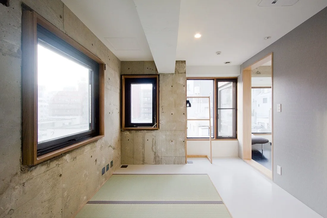 A minimalist room features raw exposed concrete walls, a green tatami mat inlaid in the floor, and large windows framed in warm wood.