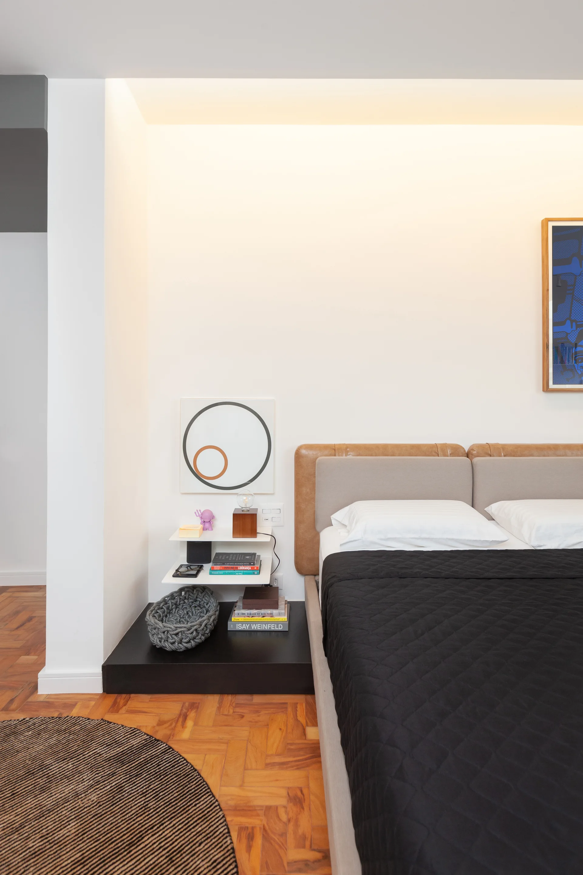 A minimalist bedroom featuring a bed with a tan leather and beige headboard, a white tiered floating nightstand, and herringbone parquet floors.