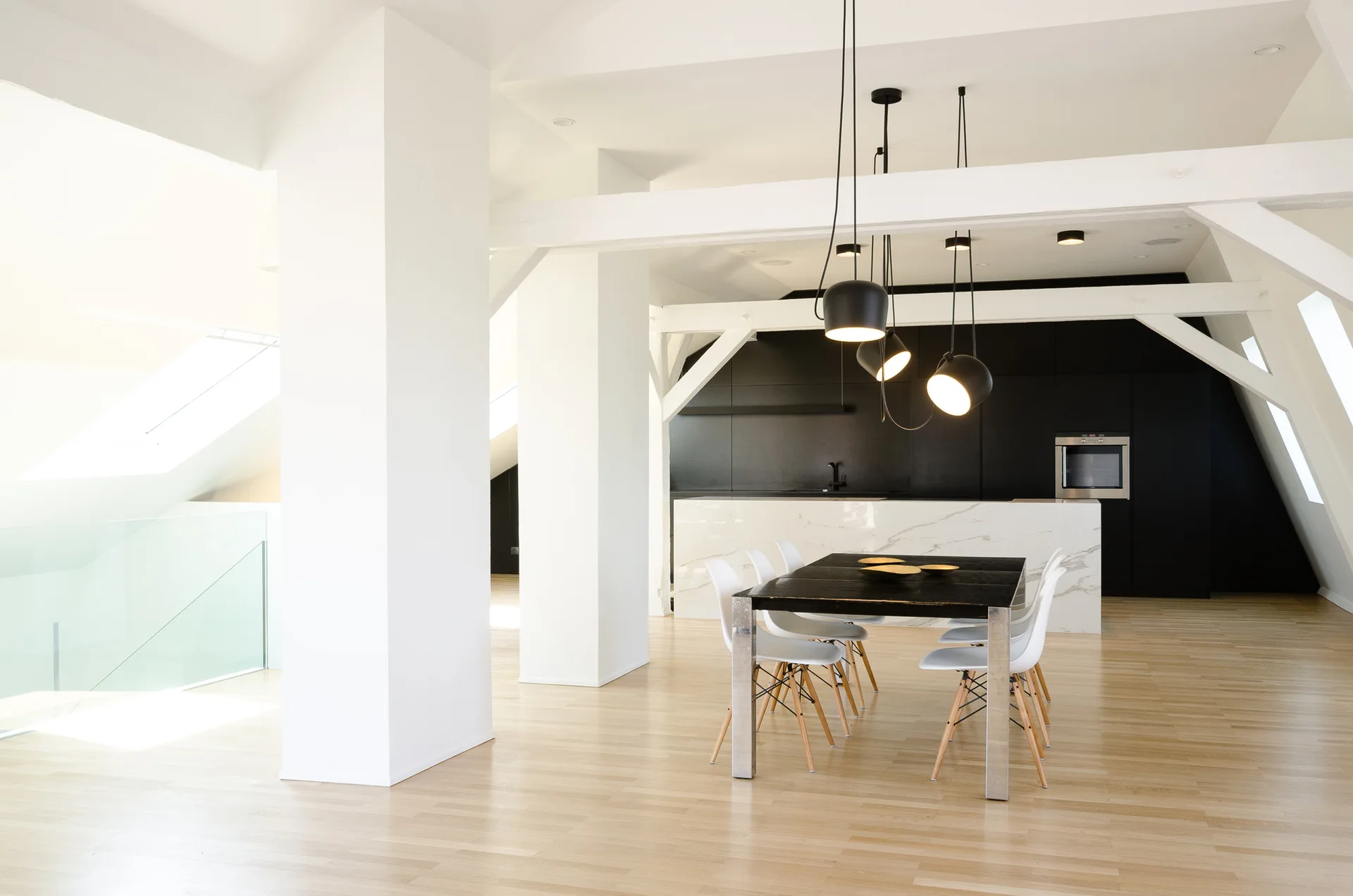 A minimalist attic dining room features a black table, white chairs, and a cluster of black pendant lights beneath exposed white ceiling beams.