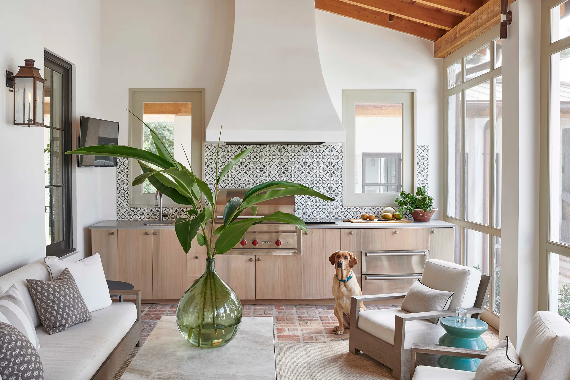 A covered porch featuring an outdoor kitchen with light wood cabinets, a patterned tile backsplash, and a seating area on a brick floor.